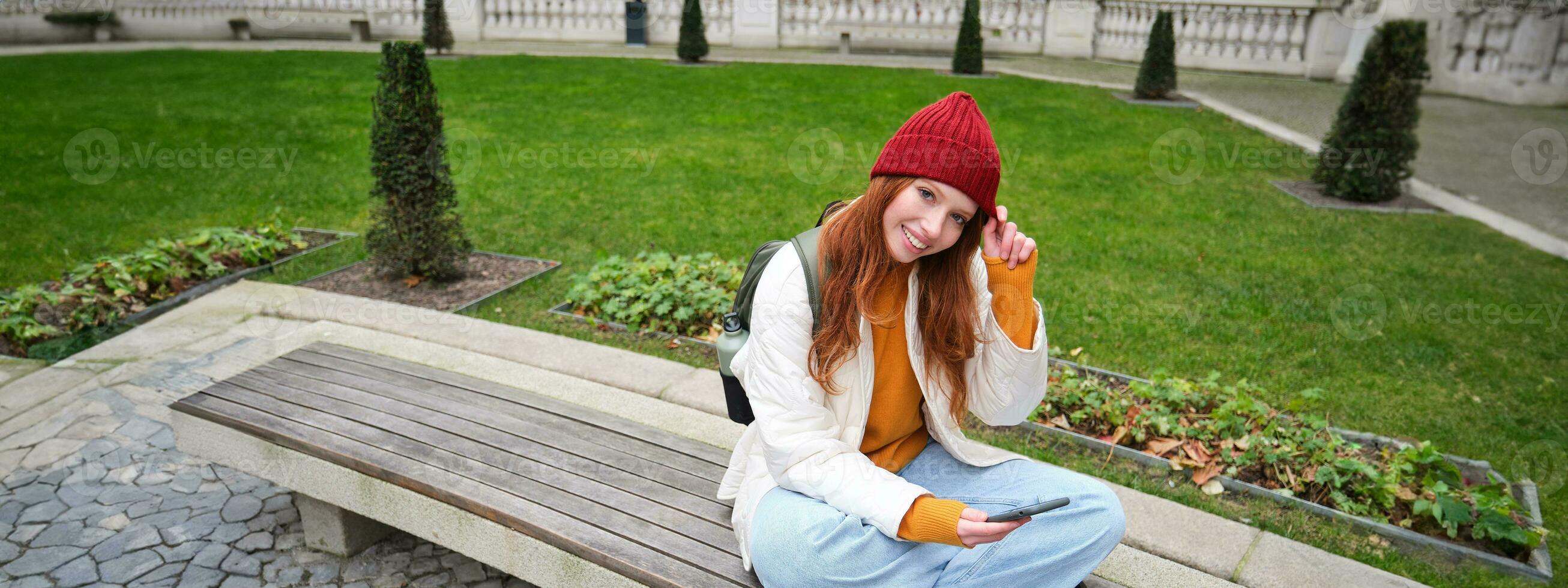 Young smiling redhead girl sits on bench and uses smartphone app, reads news online, watches video on mobile phone while relaxed in park photo