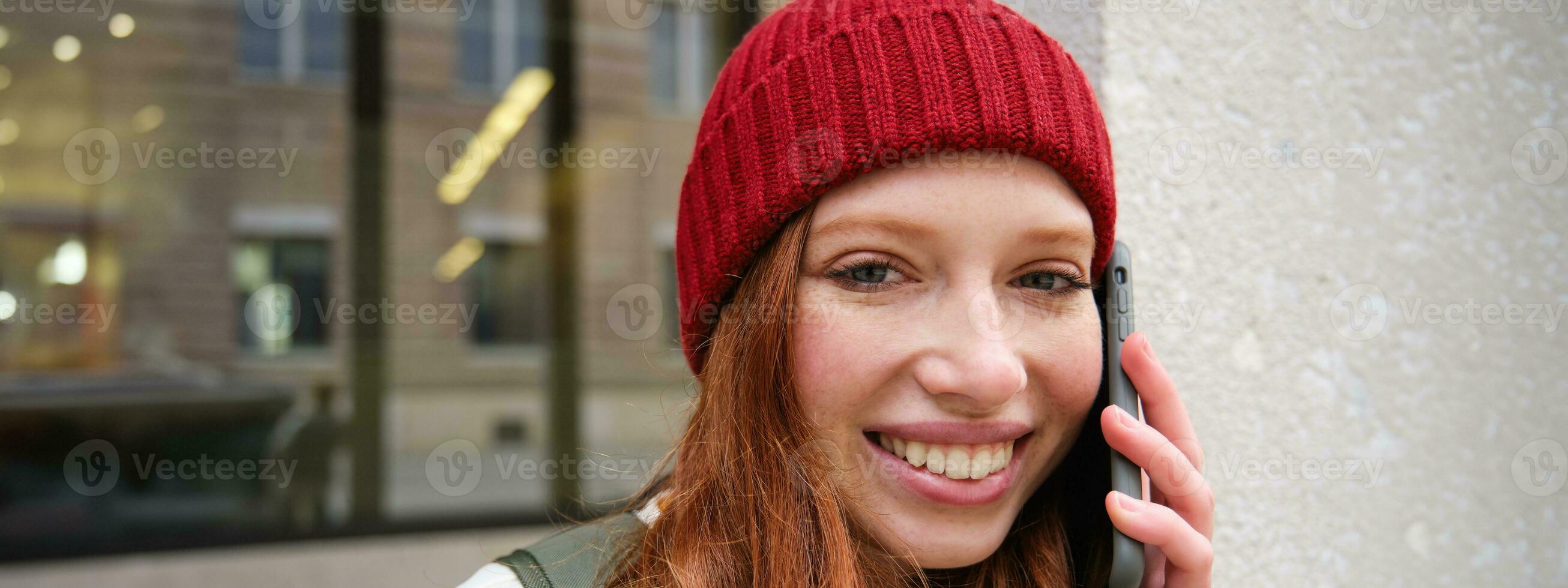 Young people and mobile connection. Happy redhead girl talks on phone, makes telephone call, stands outdoors with backpack and uses smartphone app photo