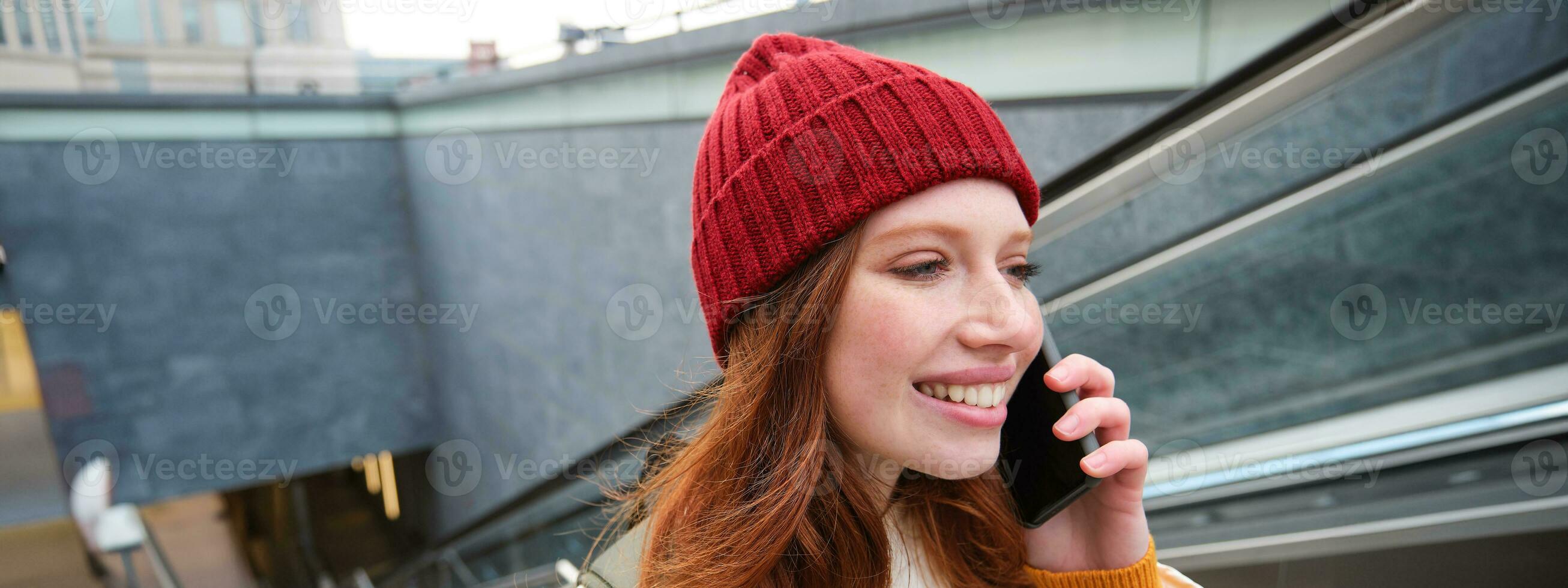 Portrait of happy redhead woman walking around town with smartphone, calling someone, talking on mobile phone outdoors photo