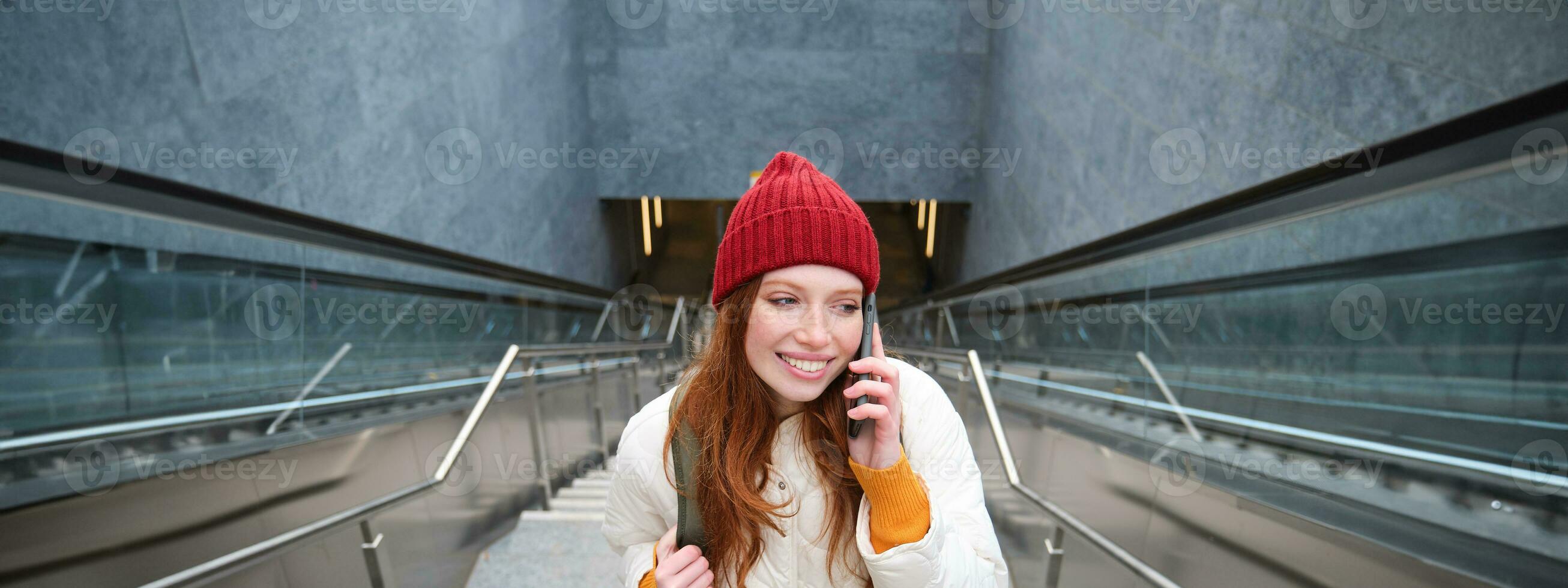 retrato de contento pelirrojo mujer caminando alrededor pueblo con teléfono inteligente, vocación alguien, hablando en móvil teléfono al aire libre foto