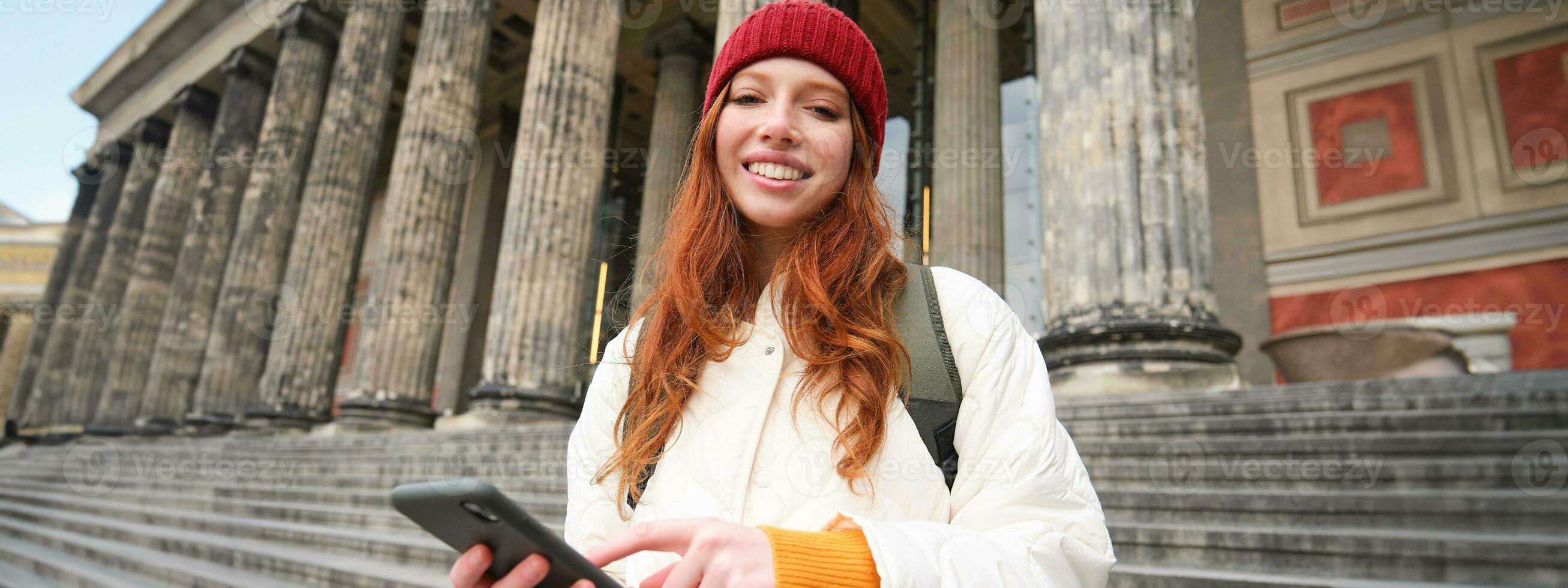 Portrait of young urban girl with backpack, using smartphone, walking around city, looking at map on mobile app, checking direction photo