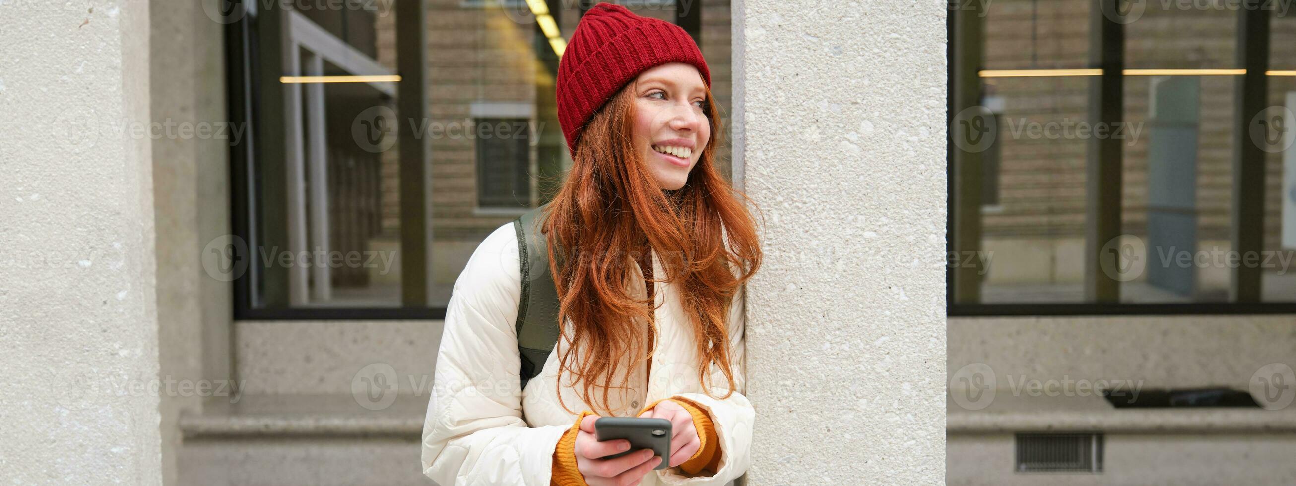 Beautiful redhead girl with smartphone, standing on street, holding mobile phone, using taxi app, connects to public wifi and uses internet, sends a text message photo