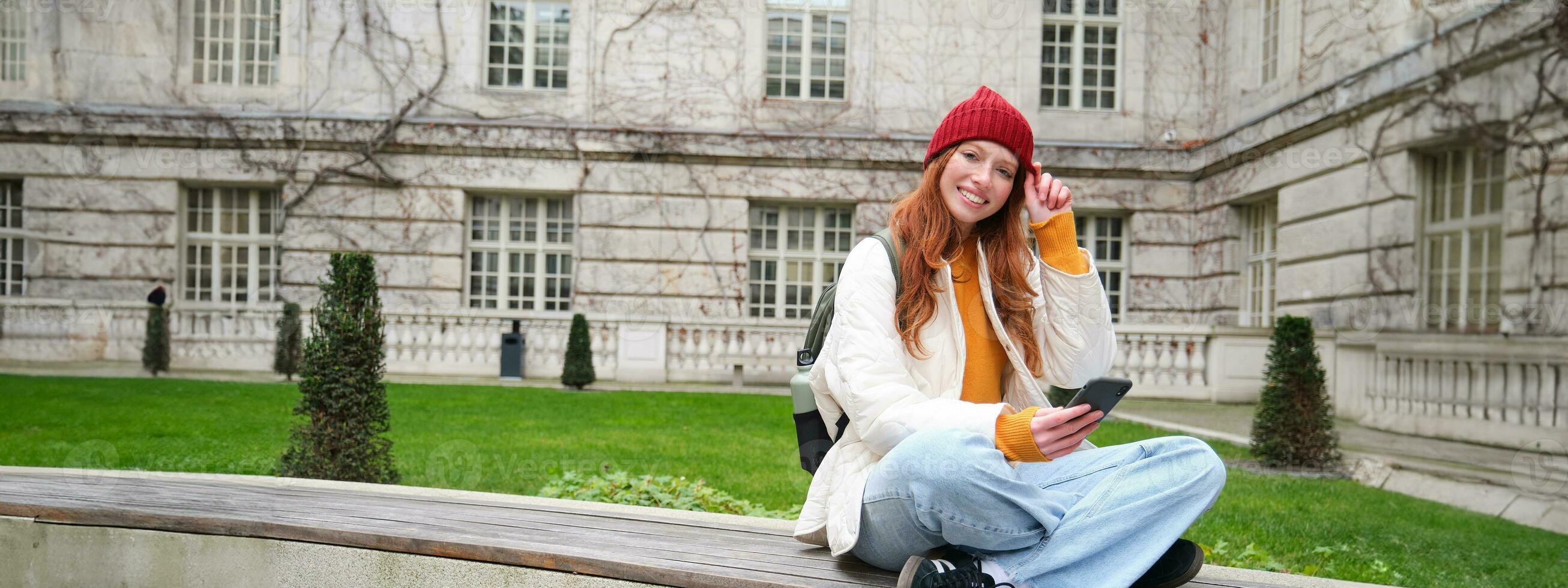 Young smiling redhead girl sits on bench and uses smartphone app, reads news online, watches video on mobile phone while relaxed in park photo
