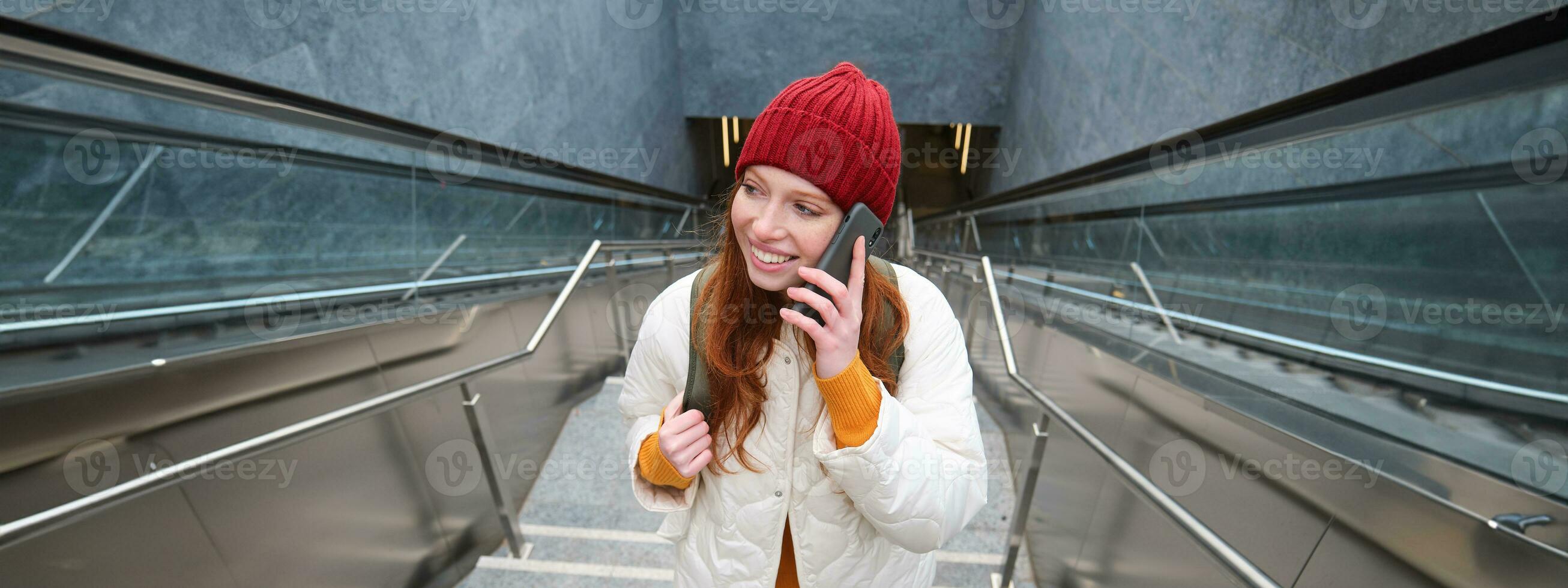 retrato de joven pelirrojo mujer camina alrededor ciudad, va arriba escalera con móvil teléfono, negociaciones en teléfono inteligente y sonrisas foto