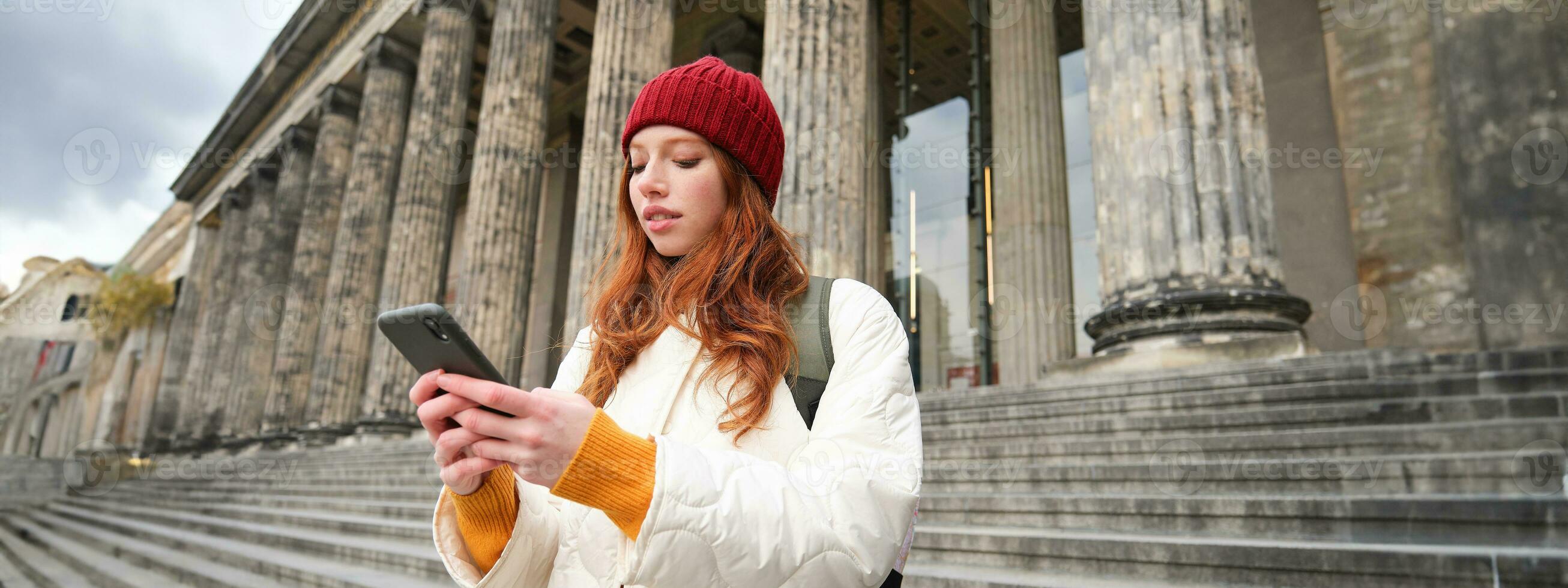 Portrait of young urban girl with backpack, using smartphone, walking around city, looking at map on mobile app, checking direction photo