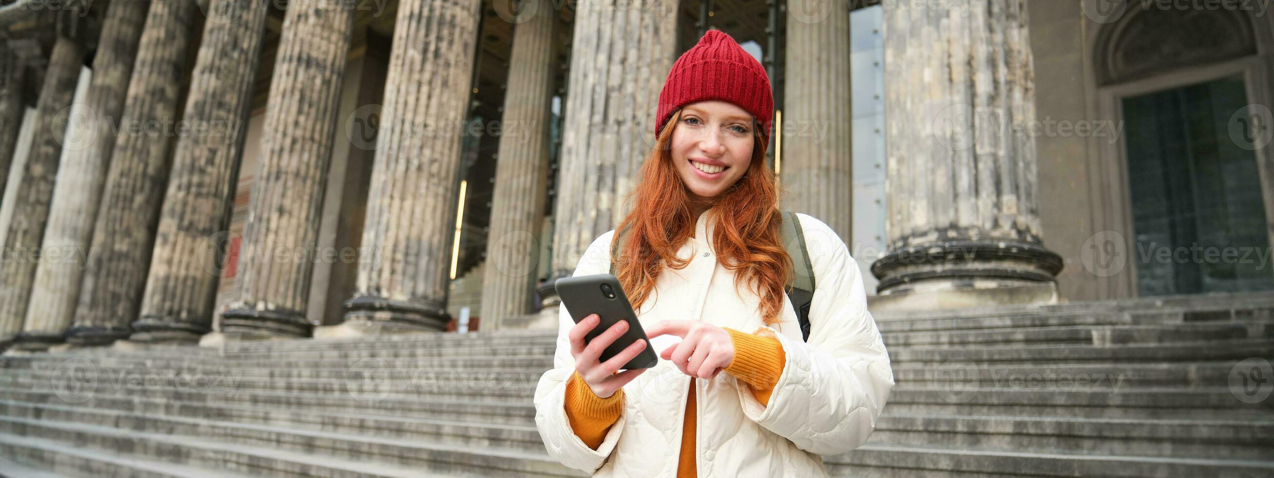 Portrait of young redhead woman holding mobile phone, using smartphone app, walking in city centre, using map application or texting a message photo