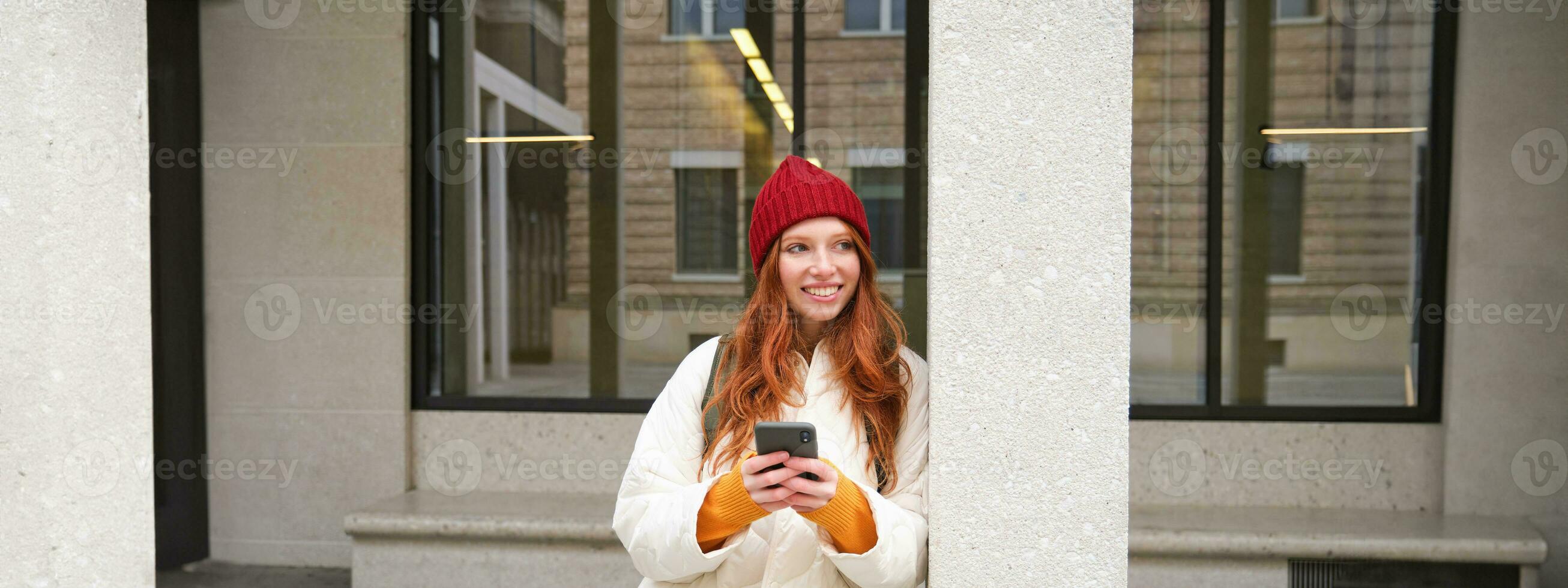 Beautiful redhead girl with smartphone, standing on street, holding mobile phone, using taxi app, connects to public wifi and uses internet, sends a text message photo