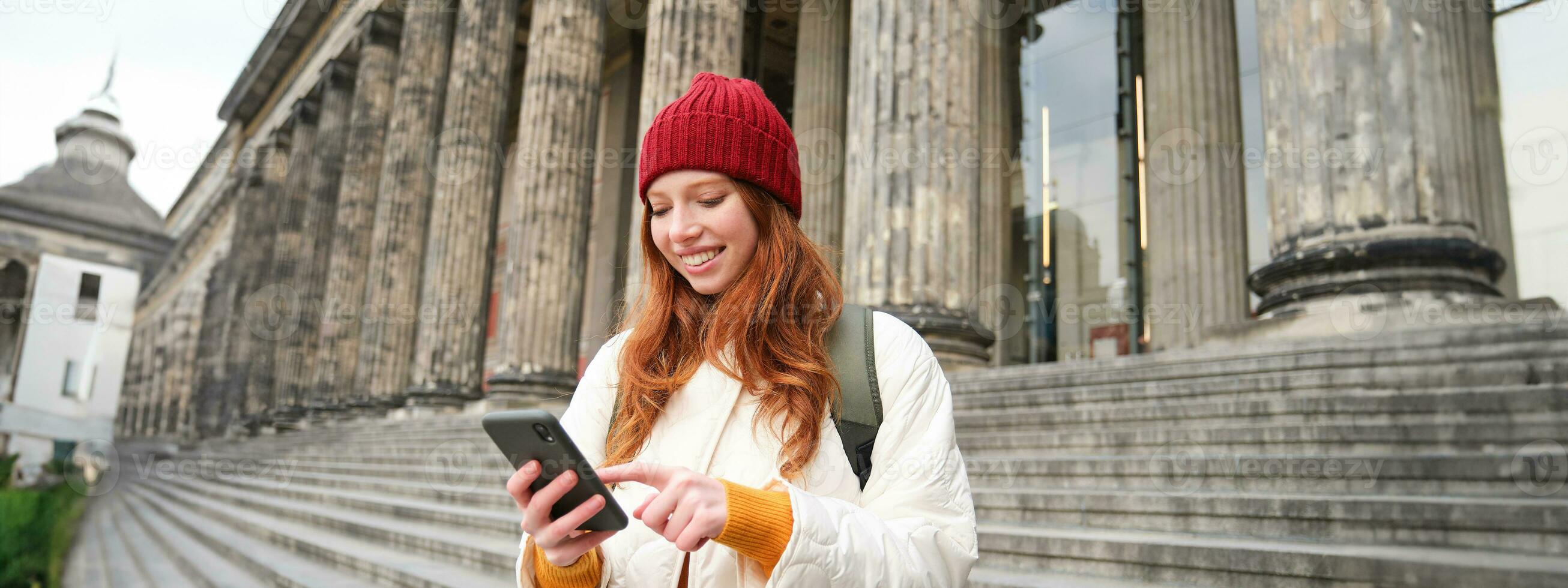 Portrait of young urban girl with backpack, using smartphone, walking around city, looking at map on mobile app, checking direction photo