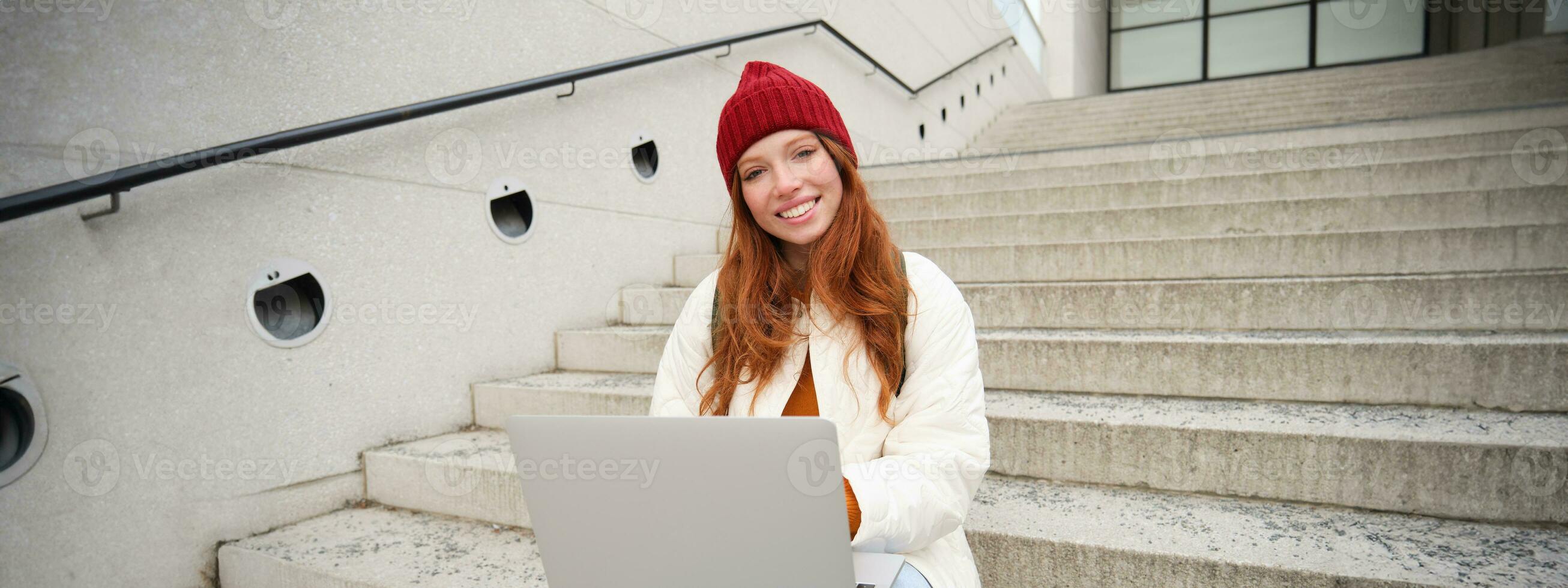 Smiling redhead girl, student sits on stairs outdoors and uses laptop, connects to public wifi in city and works on project, uses internet on computer photo
