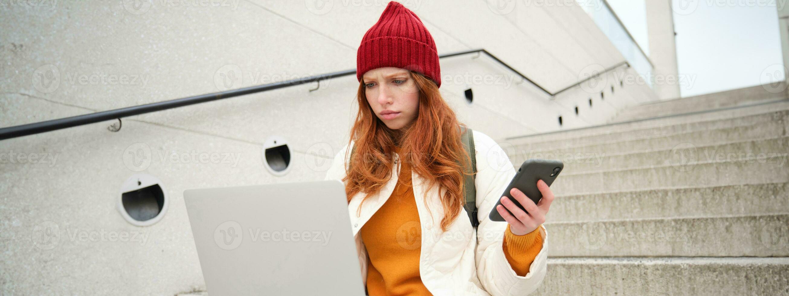 retrato de pelirrojo mujer se sienta en escaleras, usos ordenador portátil y sostiene teléfono inteligente, mira confuso y trastornado a computadora pantalla, intentos a conectar a Wifi público Internet foto