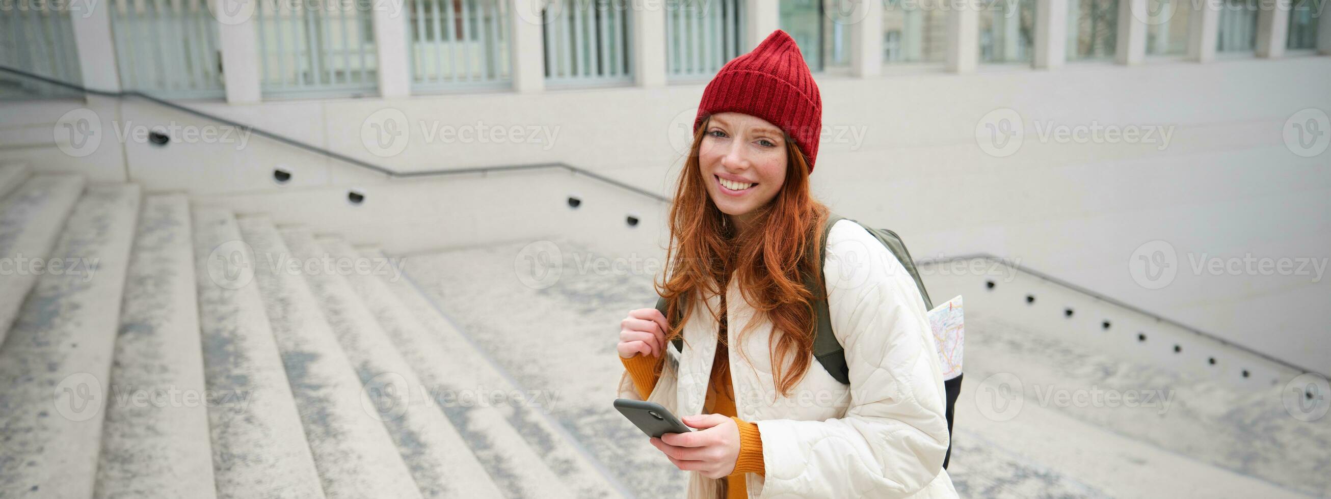 Smiling redhead girl, student tourist walks around city, goes up the stairs, looks at mobile phone map to get around town, sends message on smartphone photo