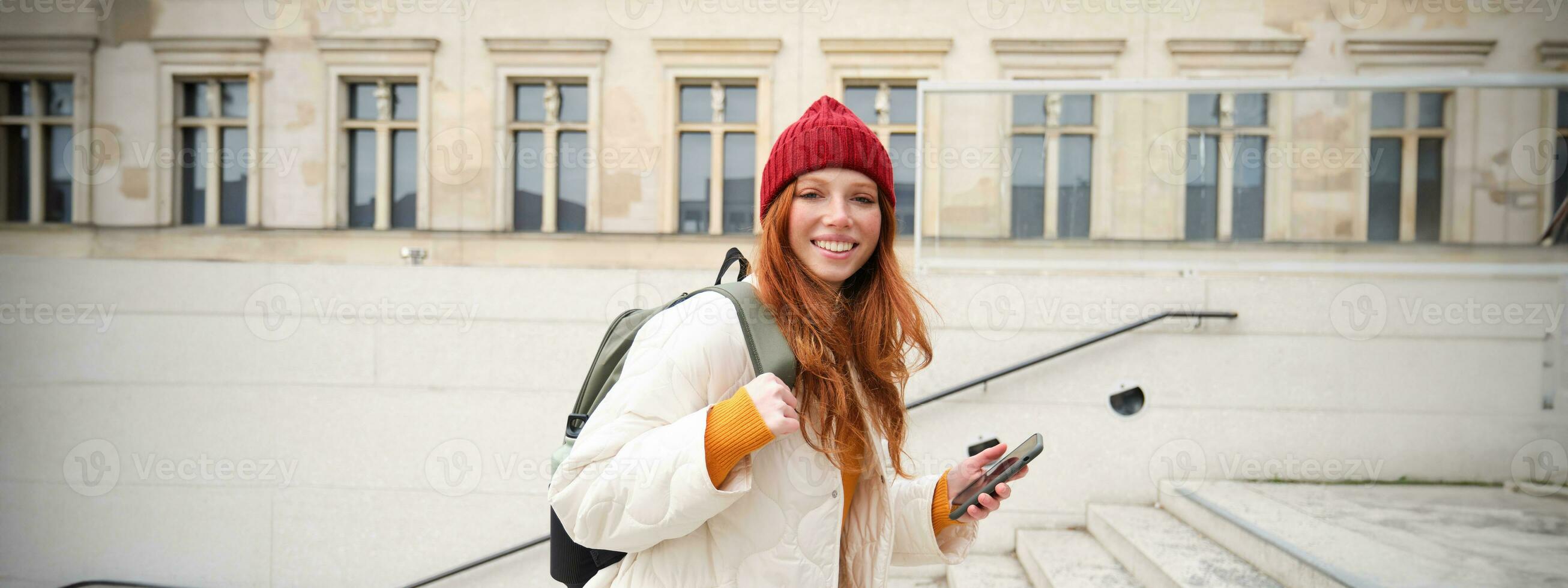 Smiling redhead girl, student tourist walks around city, goes up the stairs, looks at mobile phone map to get around town, sends message on smartphone photo