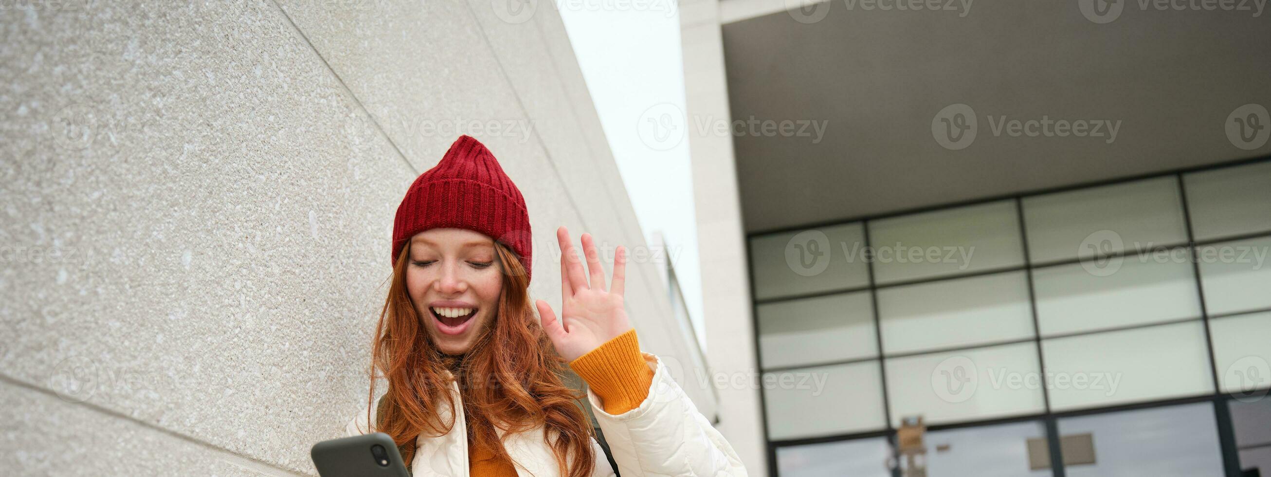 Vertical shot of girl waves at mobile phone, stands on street stairs and waves hand at smartphone camera, video chats, says hello, talks to friend on telephone photo