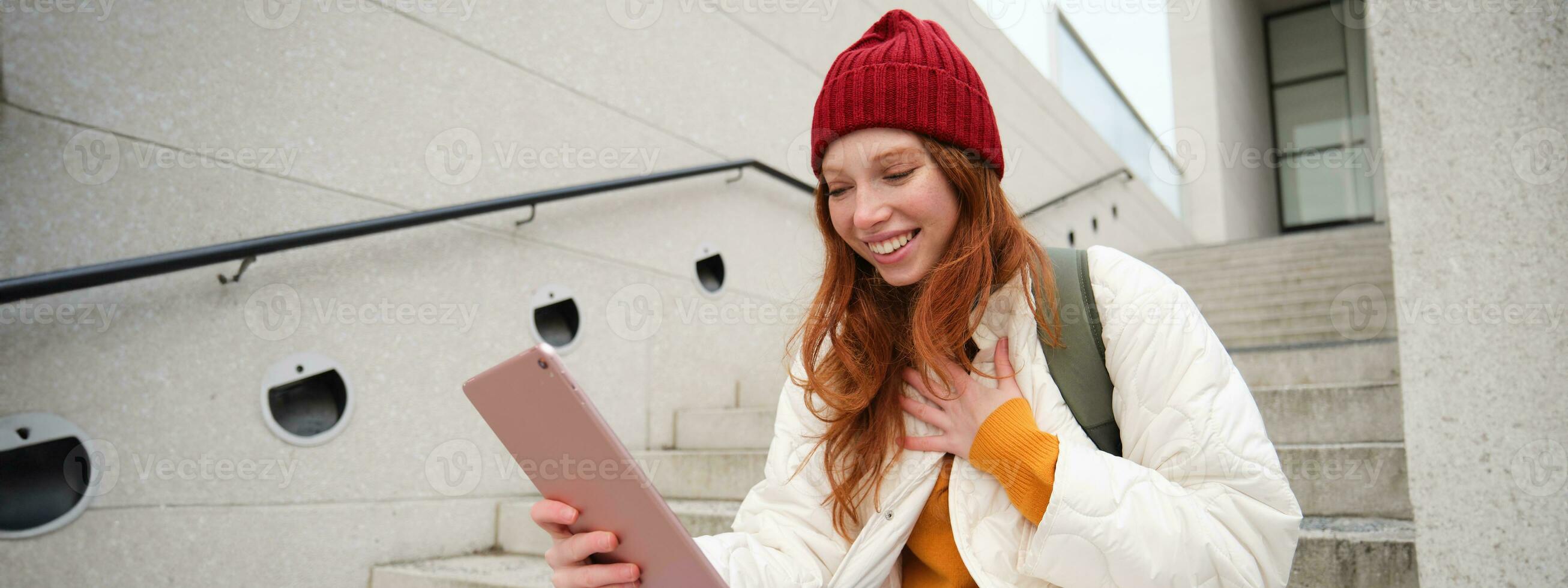 Happy stylish redhead girl, student in red hat, holds digital tablet, uses social media app, searches something online, connects to wifi photo