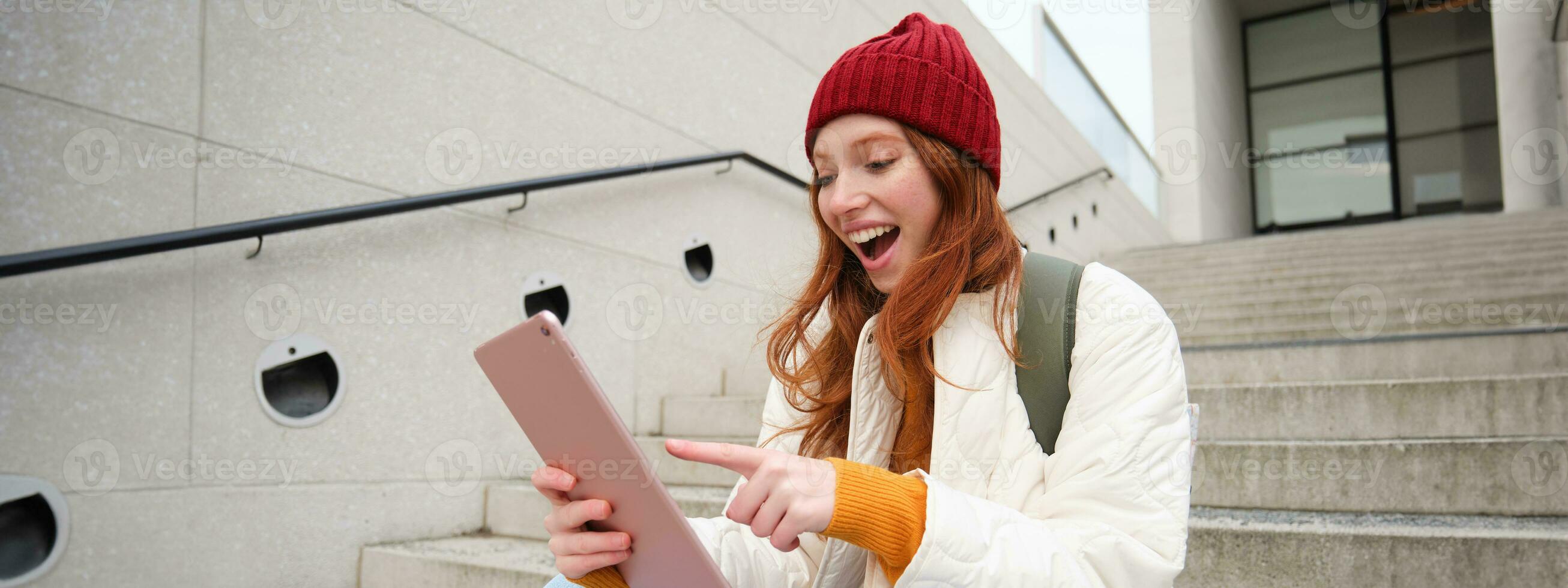 Young stylish girl, redhead female students sits on stairs outdoors with digital tablet, reads, uses social media app on gadget, plays games while waits on street photo