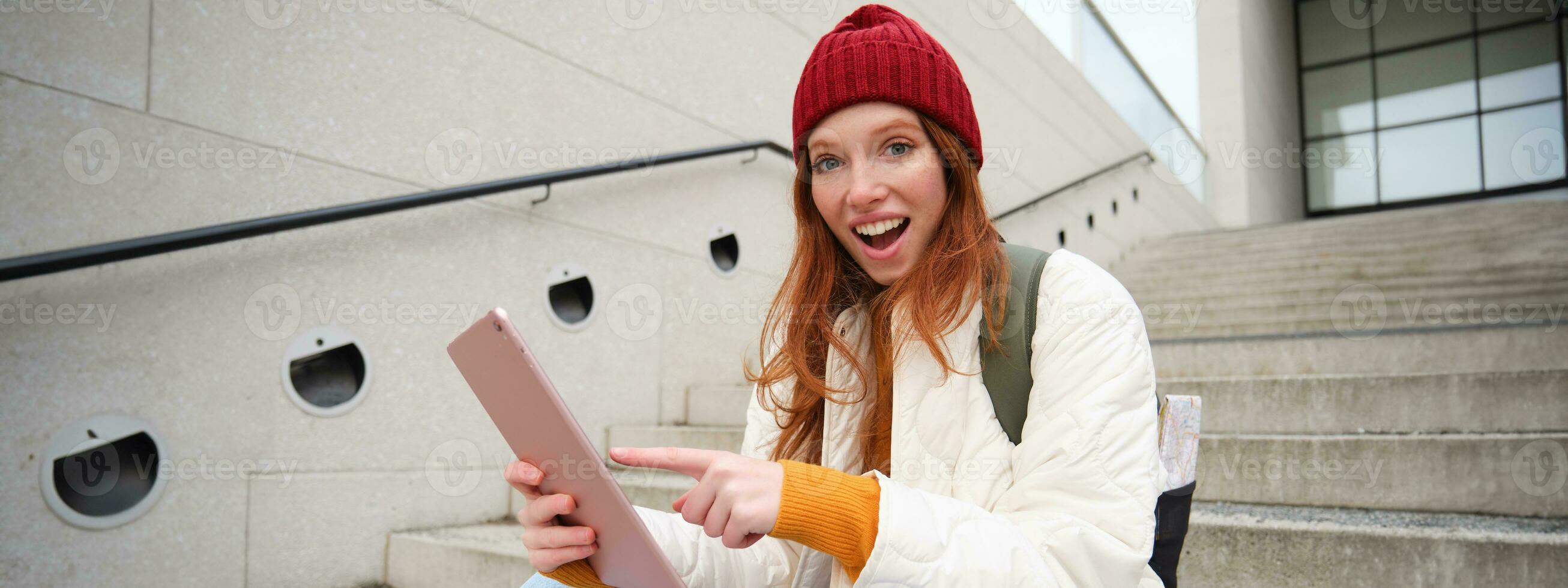 Young stylish girl, redhead female students sits on stairs outdoors with digital tablet, reads, uses social media app on gadget, plays games while waits on street photo