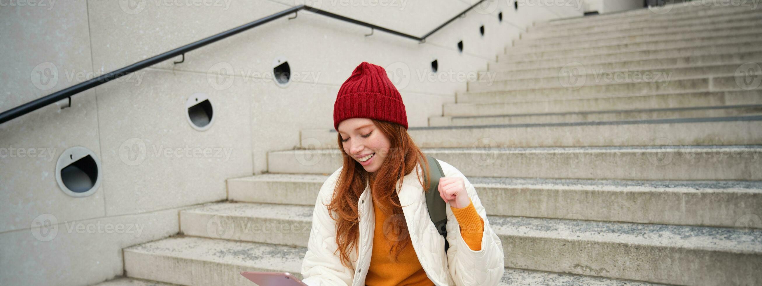 Young beautiful girl with long red hair, smiles, uses internet application, connect to wifi near campus, sits on stairs with digital tablet photo