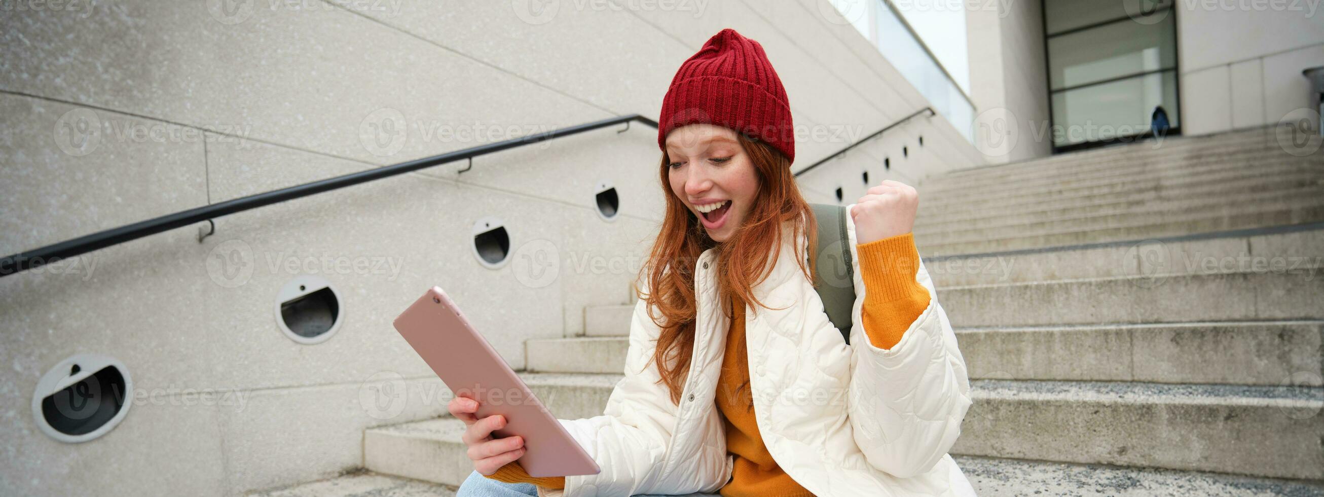 contento elegante pelirrojo chica, estudiante en rojo sombrero, sostiene digital tableta, usos social medios de comunicación aplicación, búsquedas alguna cosa en línea, conecta a Wifi foto