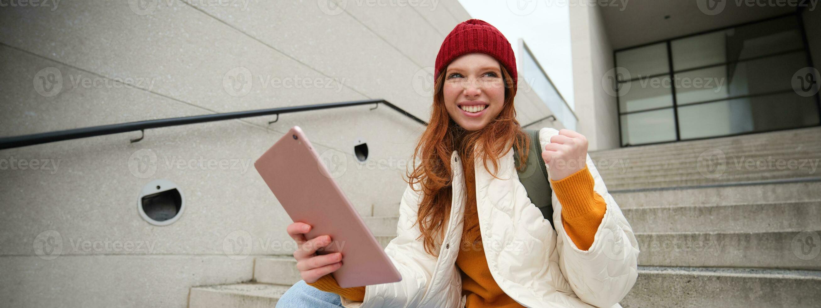 Happy stylish redhead girl, student in red hat, holds digital tablet, uses social media app, searches something online, connects to wifi photo