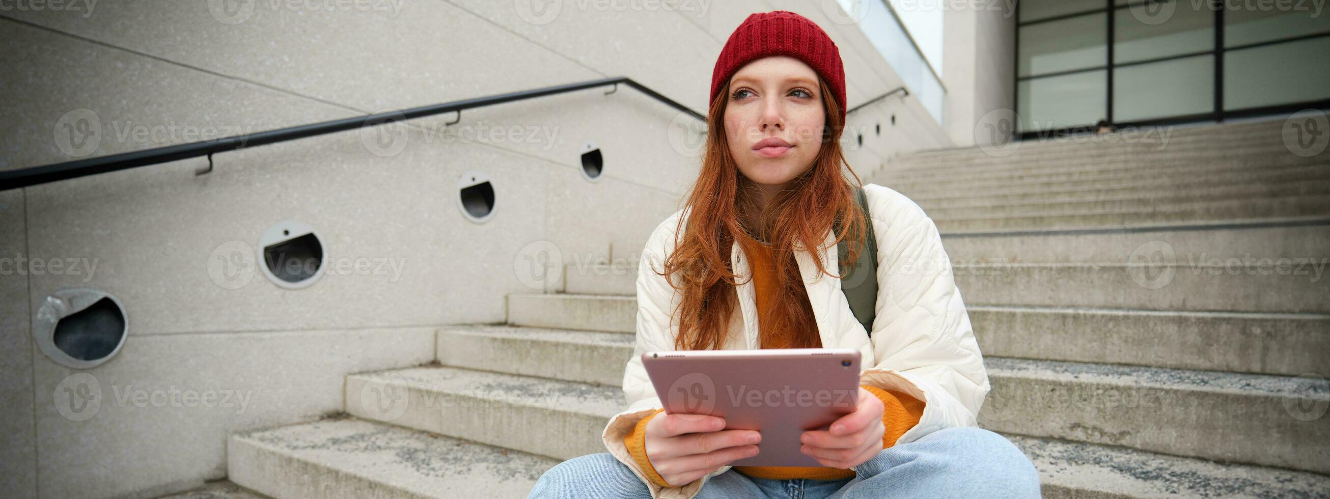 Young stylish girl, redhead female students sits on stairs outdoors with digital tablet, reads, uses social media app on gadget, plays games while waits on street photo