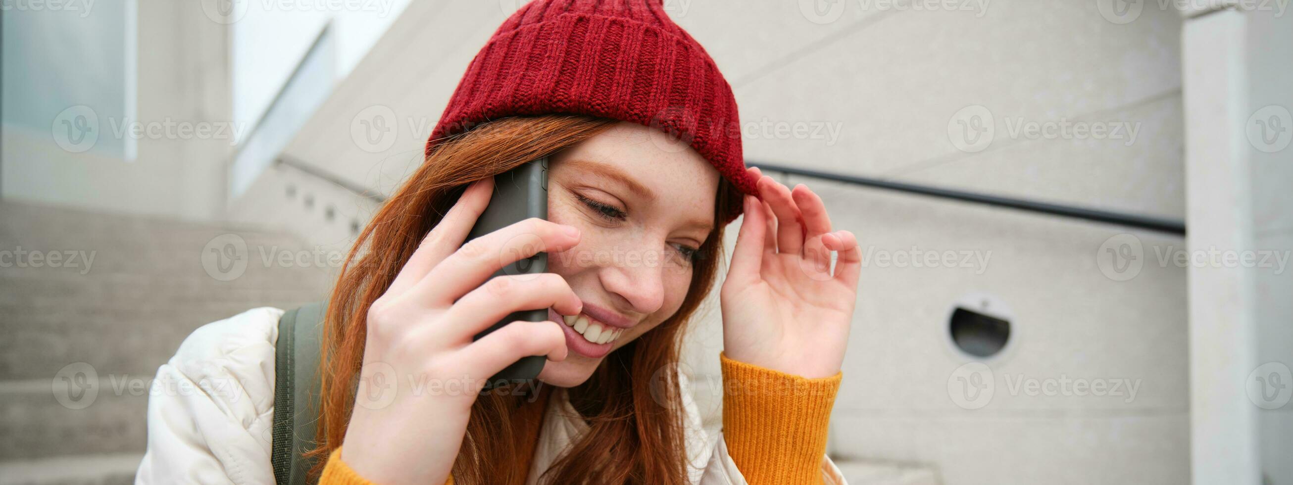 Young stylish redhead girl in red hat, sits on street and talks on mobile phone, has telephone conversation, rings her friend while relaxes outdoors photo