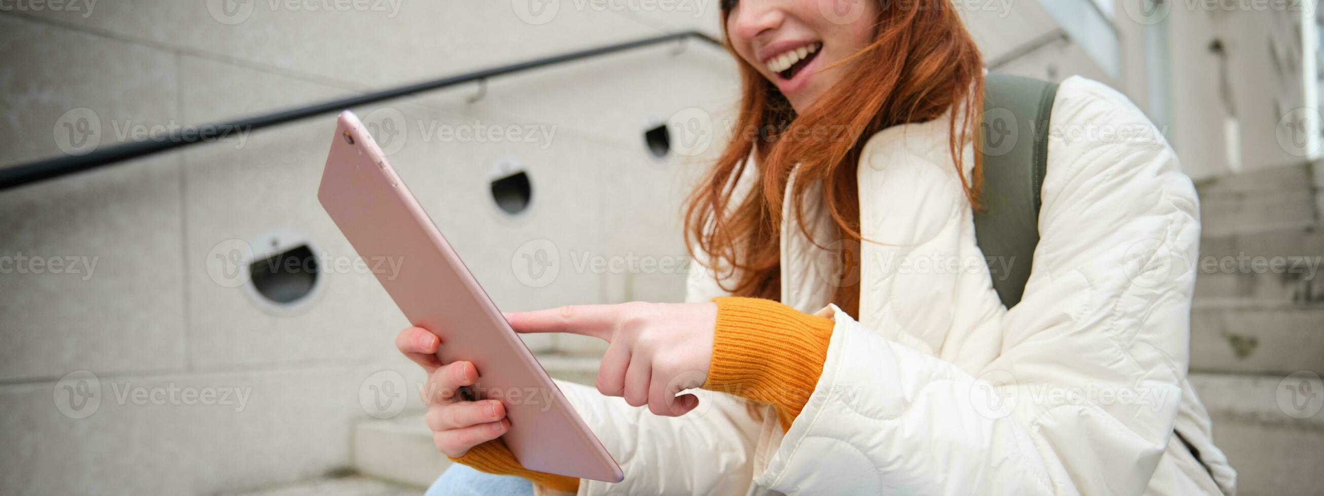 Young stylish girl, redhead female students sits on stairs outdoors with digital tablet, reads, uses social media app on gadget, plays games while waits on street photo