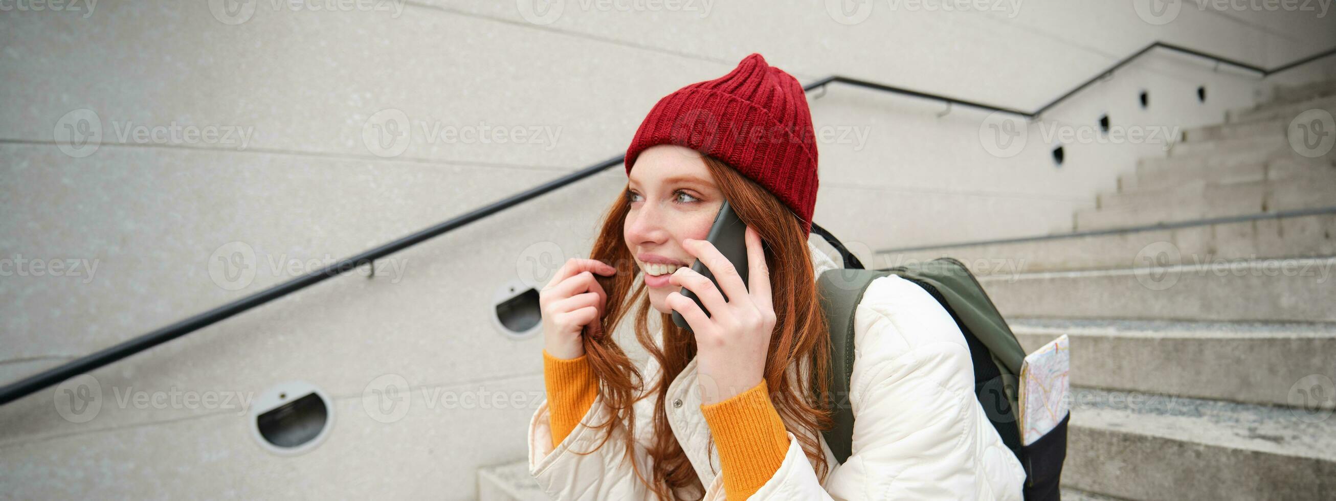 hermosa sonriente pelirrojo hembra modelo, se sienta en calle y negociaciones en móvil teléfono, usos teléfono inteligente aplicación a llamada en el extranjero, riendo durante teléfono conversacion foto