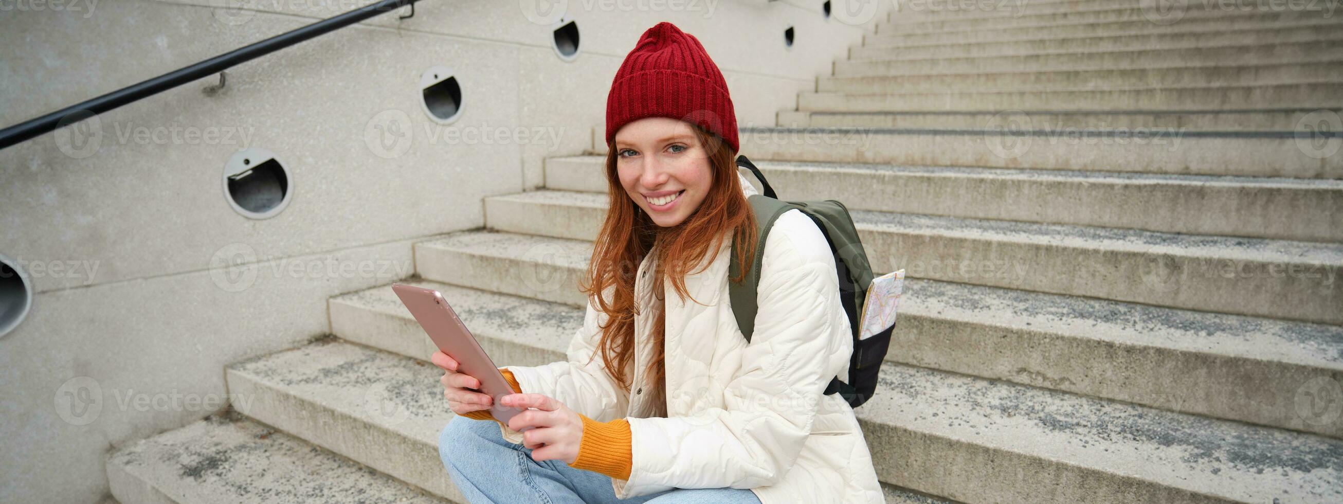 Happy stylish redhead girl, student in red hat, holds digital tablet, uses social media app, searches something online, connects to wifi photo