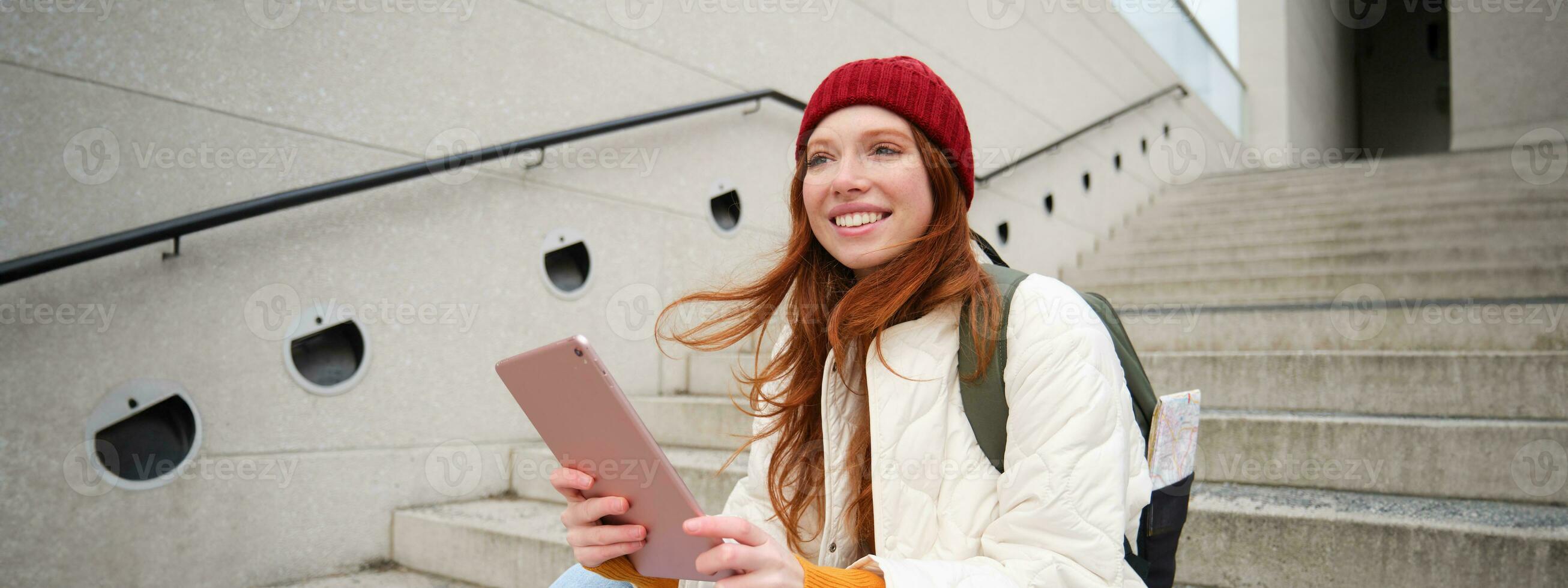 Young stylish girl, redhead female students sits on stairs outdoors with digital tablet, reads, uses social media app on gadget, plays games while waits on street photo