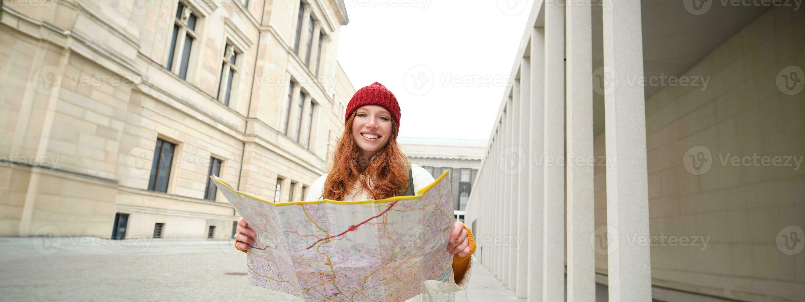 Beautiful redhead woman, tourist with city map, explores sightseeing historical landmark, walking around old town, smiling happily photo