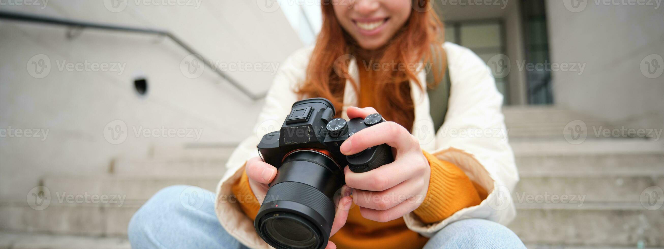 Smiling redhead girl photographer, checks her shots, holds camera and looks at screen, takes photos outdoors, walks around street and does streetstyle shooting
