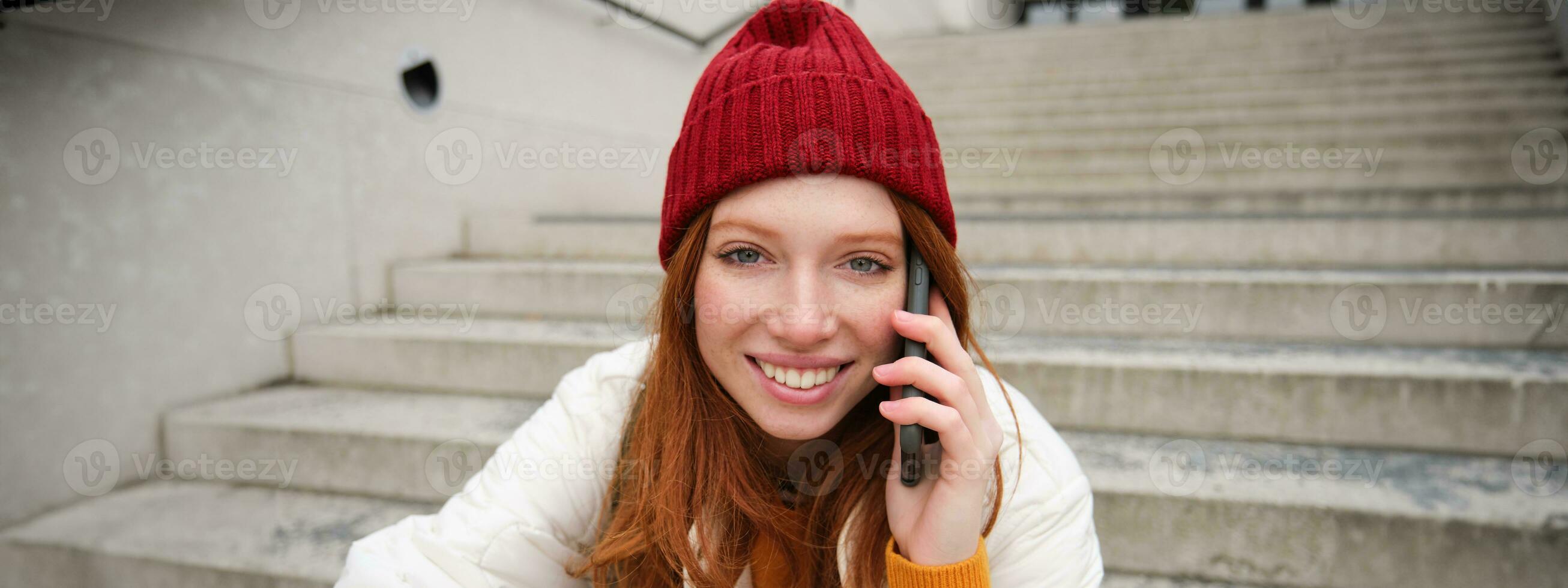 hermosa sonriente pelirrojo hembra modelo, se sienta en calle y negociaciones en móvil teléfono, usos teléfono inteligente aplicación a llamada en el extranjero, riendo durante teléfono conversacion foto