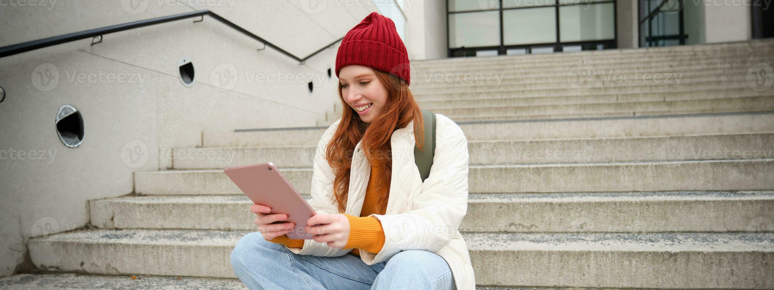 Young stylish girl, redhead female students sits on stairs outdoors with digital tablet, reads, uses social media app on gadget, plays games while waits on street photo