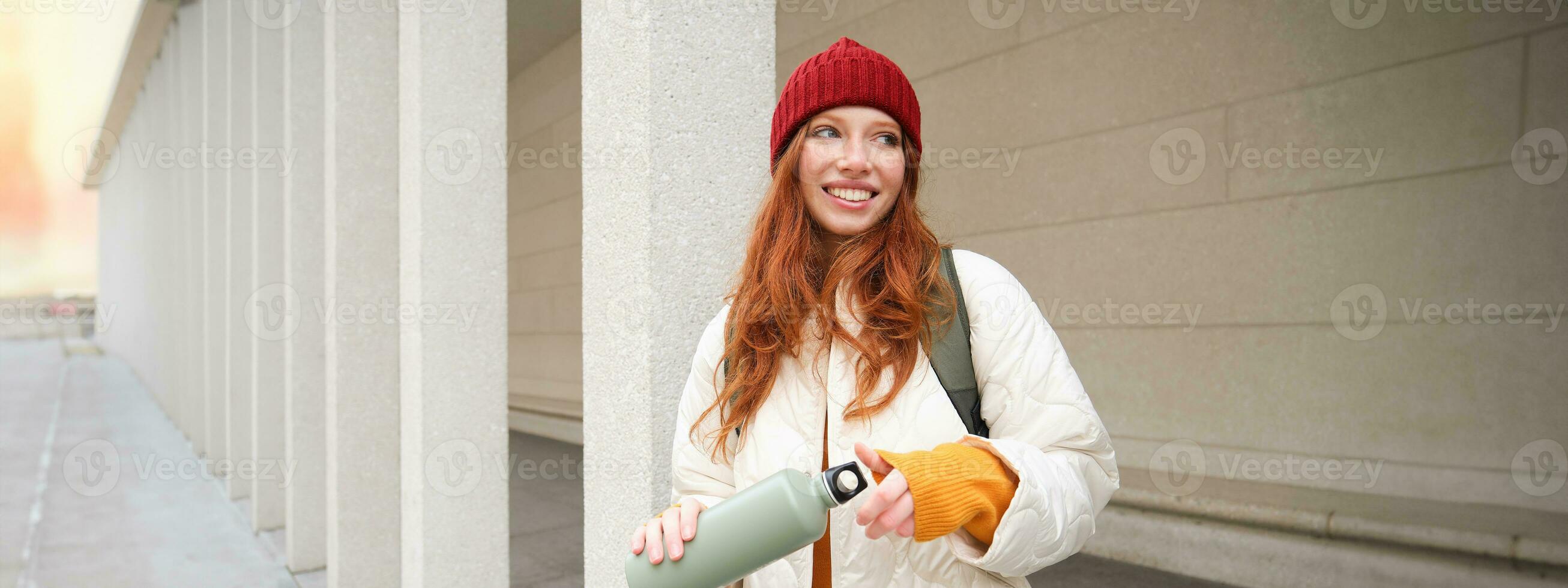 Young happy redhead woman in red hat, drinking from thermos, warming up with hot drink in her flask while walking around city, tourist relaxes with warm refreshemtn photo