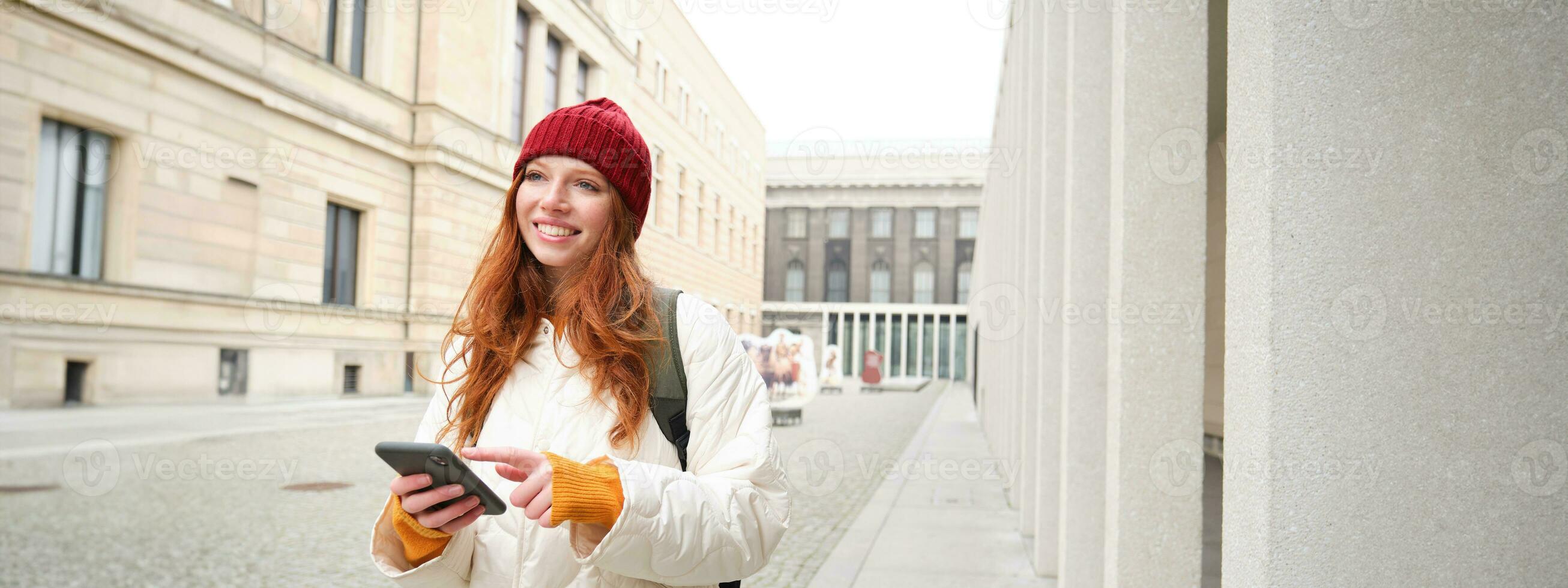 Beautiful smiling girl, tourist with backpack, holding smartphone, using map on mobile phone application, looking for sightseeing in internet app, standing outdoors photo