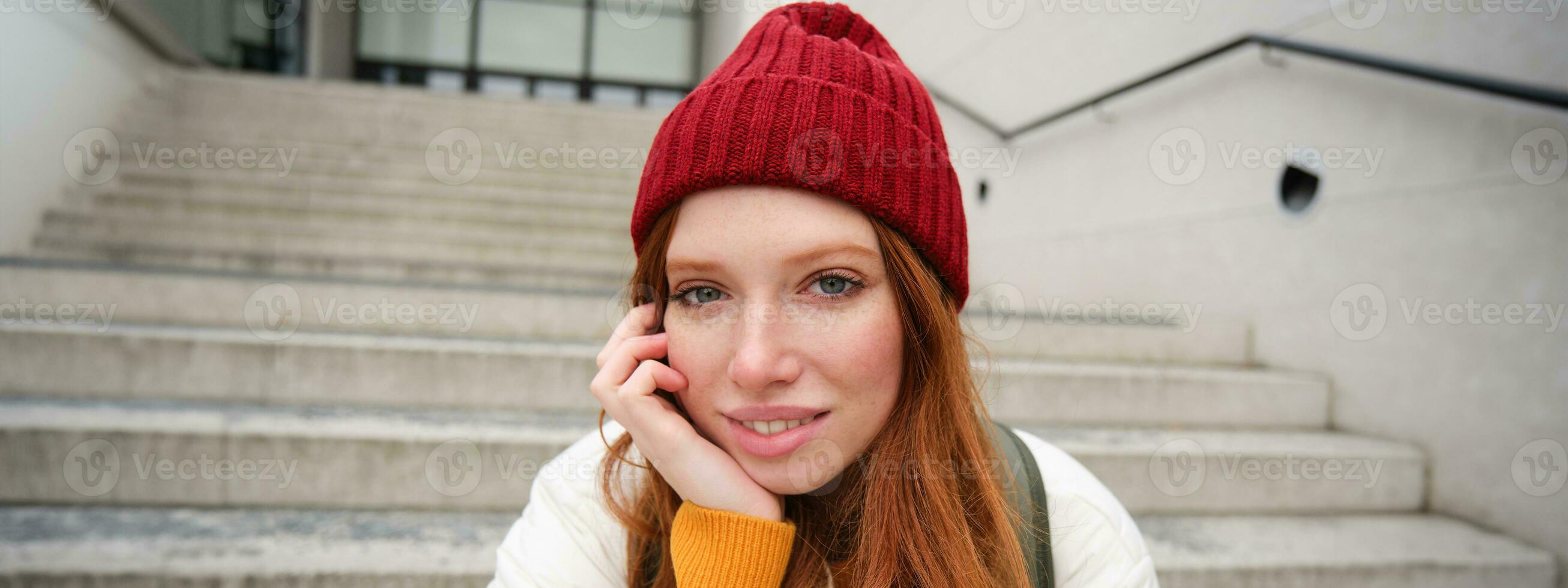 Close up portrait of beautiful redhead girl in red hat, urban woman with freckles and ginger hair, sits on stairs on street, smiles and looks gorgeous photo