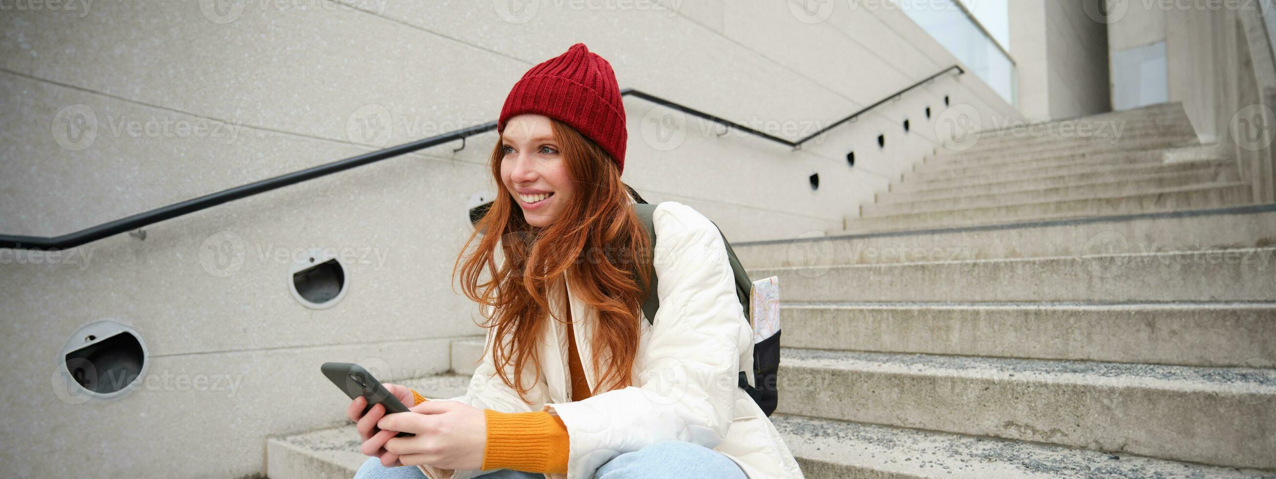 elegante europeo niña con rojo cabello, se sienta en público escalera con teléfono inteligente, lugares en línea orden, envía mensaje en móvil teléfono social aplicación, sonrisas felizmente foto