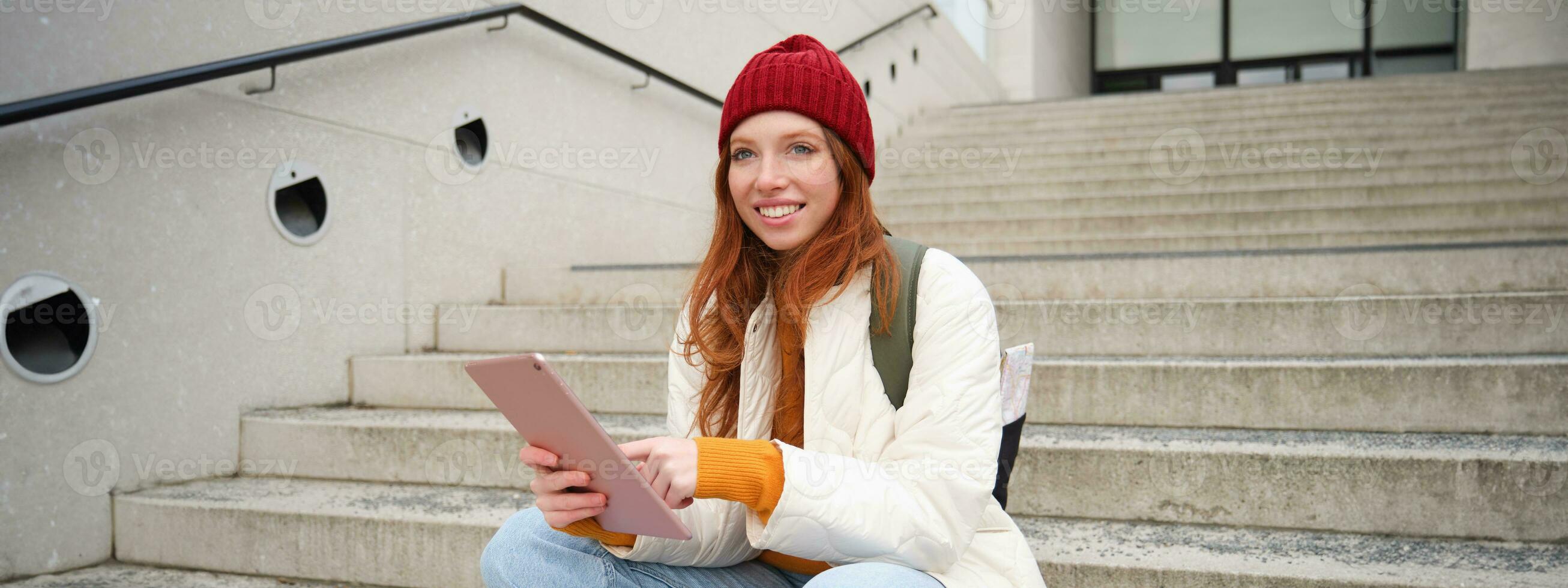 Young stylish girl, redhead female students sits on stairs outdoors with digital tablet, reads, uses social media app on gadget, plays games while waits on street photo