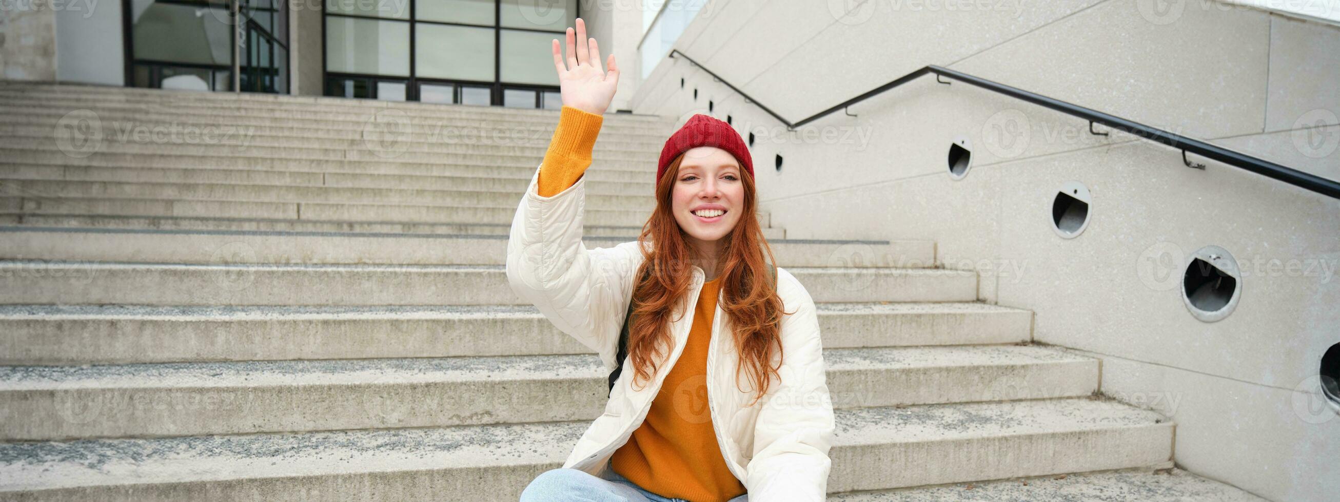 Smiling young redhead girl waits someone on stairs outdoors, waves hand at friend, holds smartphone, says hello photo