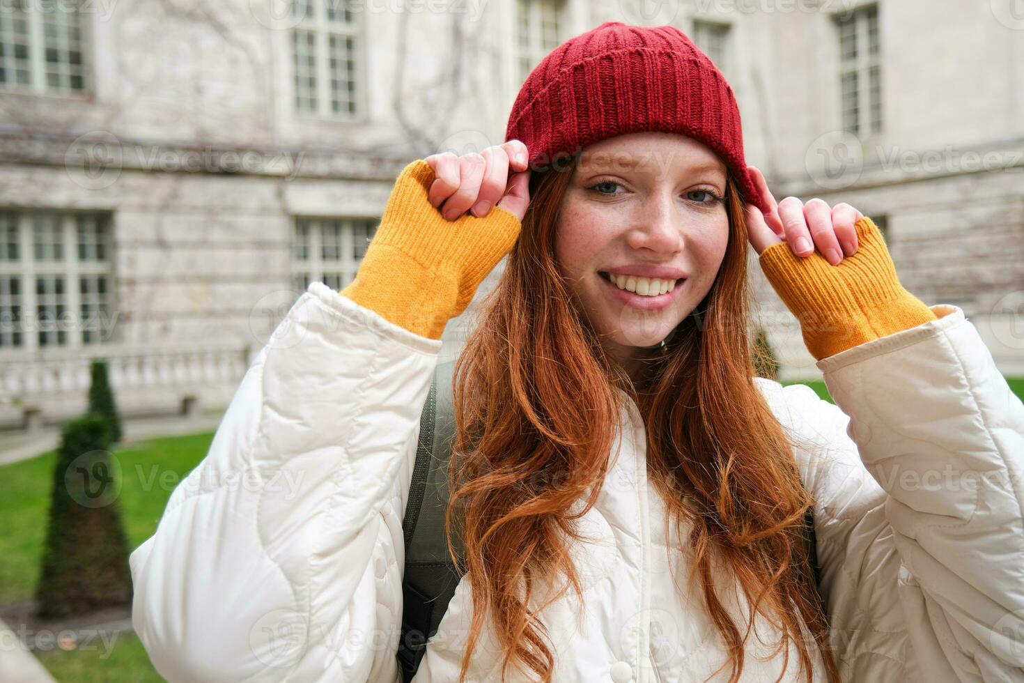 Young smiling redhead woman walking in beautiful city attractions, wearing backpack, red hat and coat, looking around with happy face photo