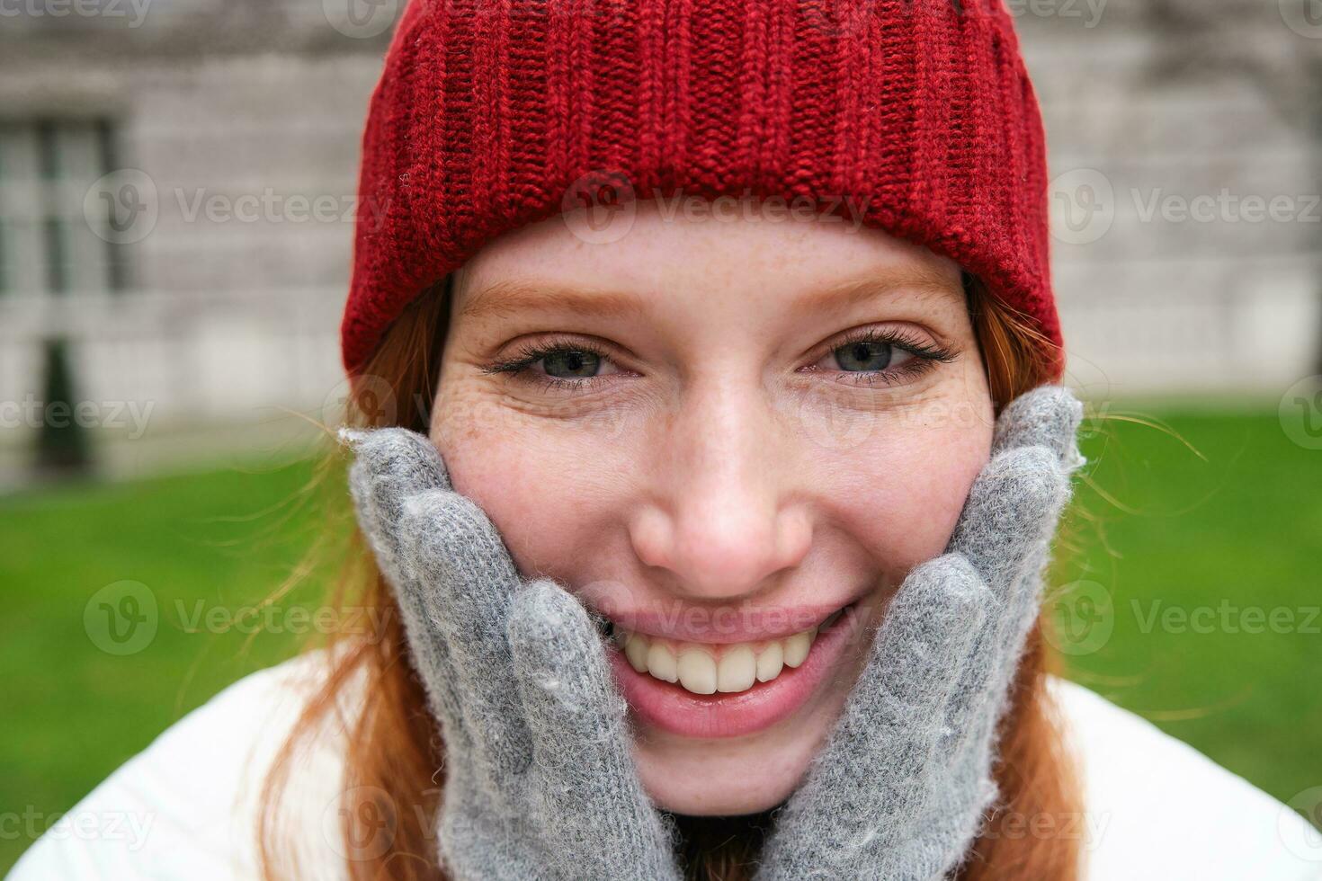 Close up portrait of beautiful redhead woman in red knitted hat, warm gloves, smiling and looking happy at camera, sitting in park photo