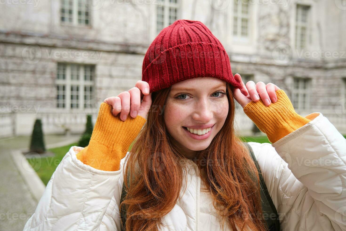 Young smiling redhead woman walking in beautiful city attractions, wearing backpack, red hat and coat, looking around with happy face photo