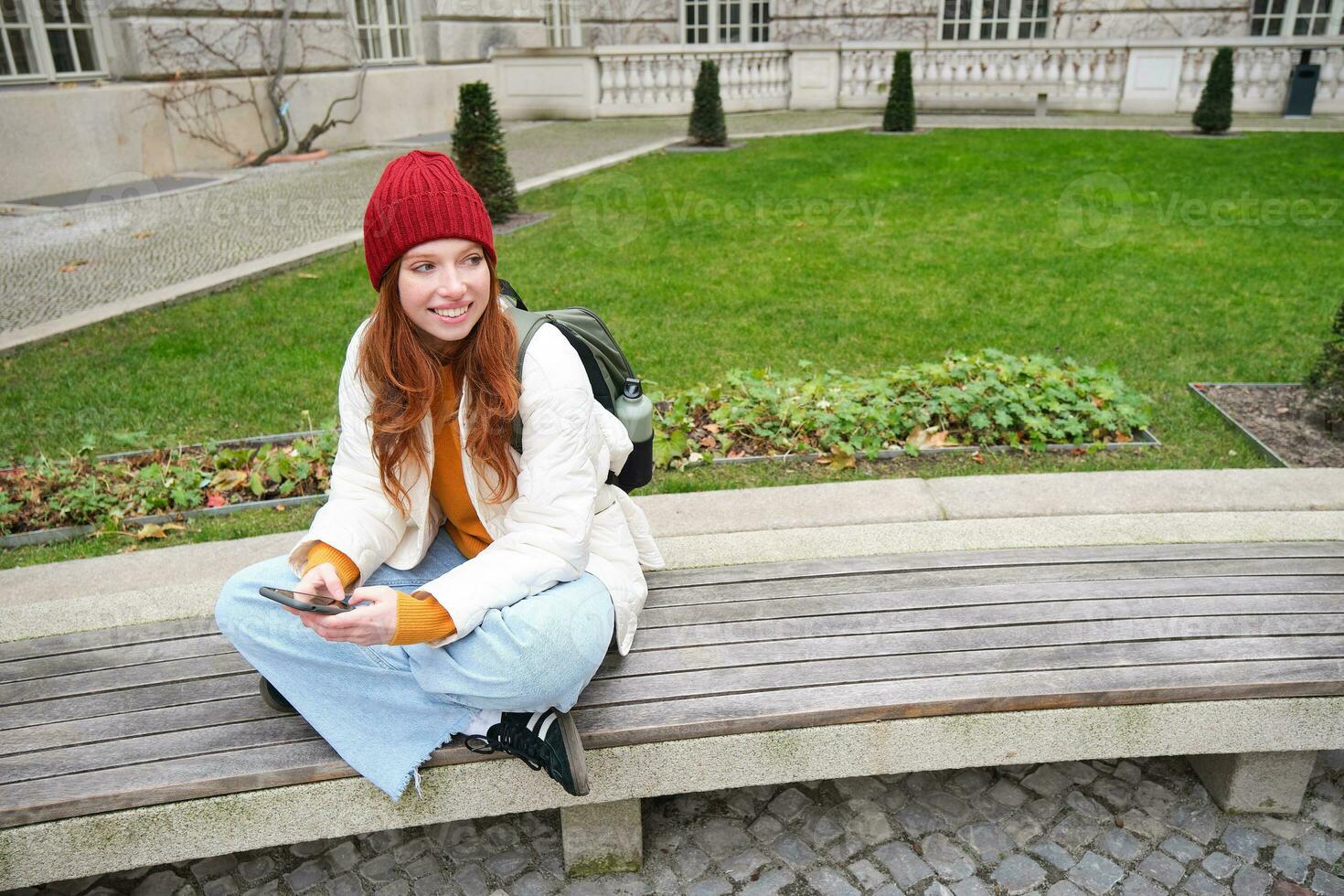 Young stylish female student with backpack, sits on bench and uses smartphone, plays on mobile phone photo