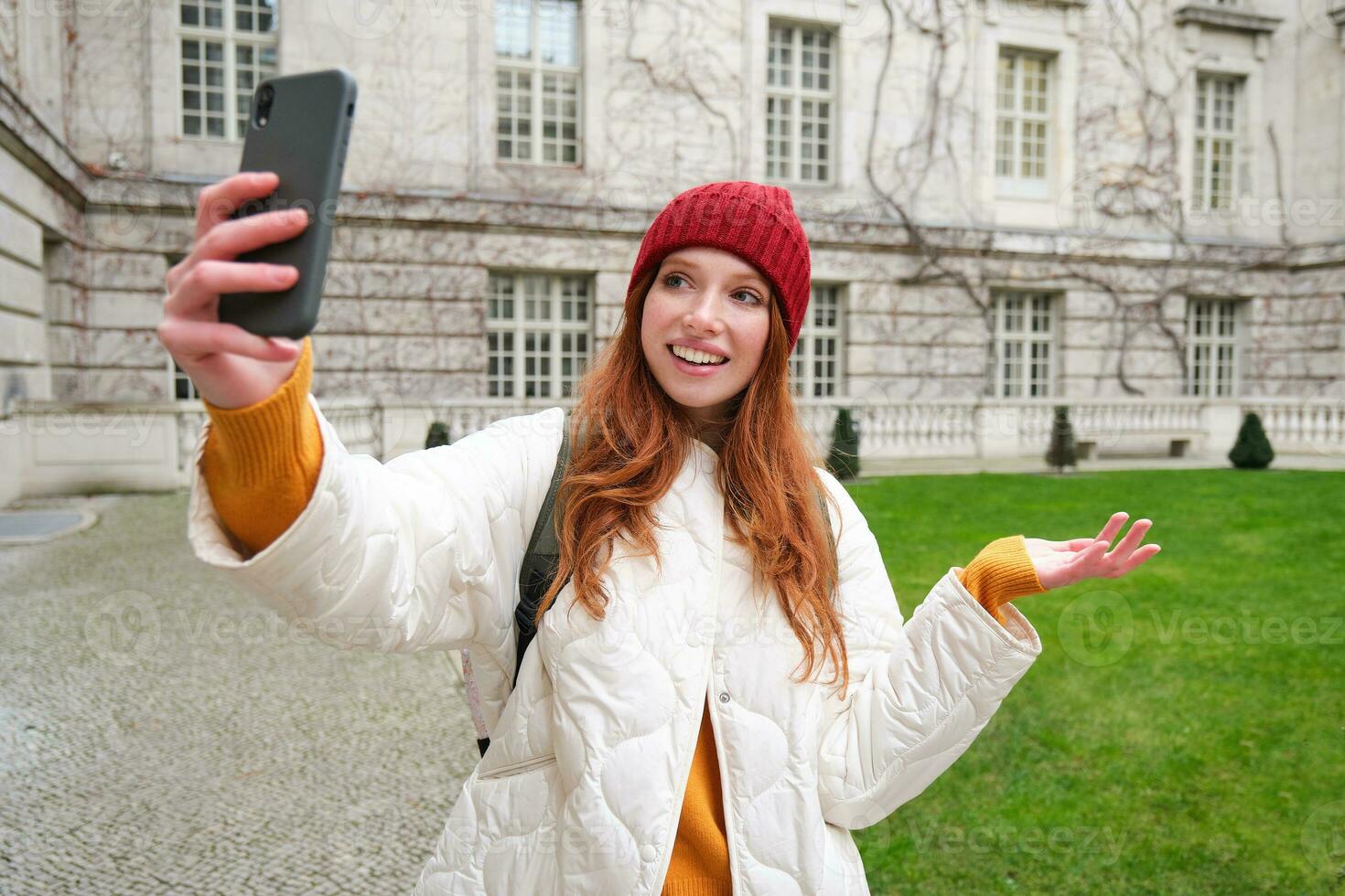 Portrait of happy girl, tourist posing near city attraction, video chats and demonstrates landmark, shows something to friend with mobile phone app photo
