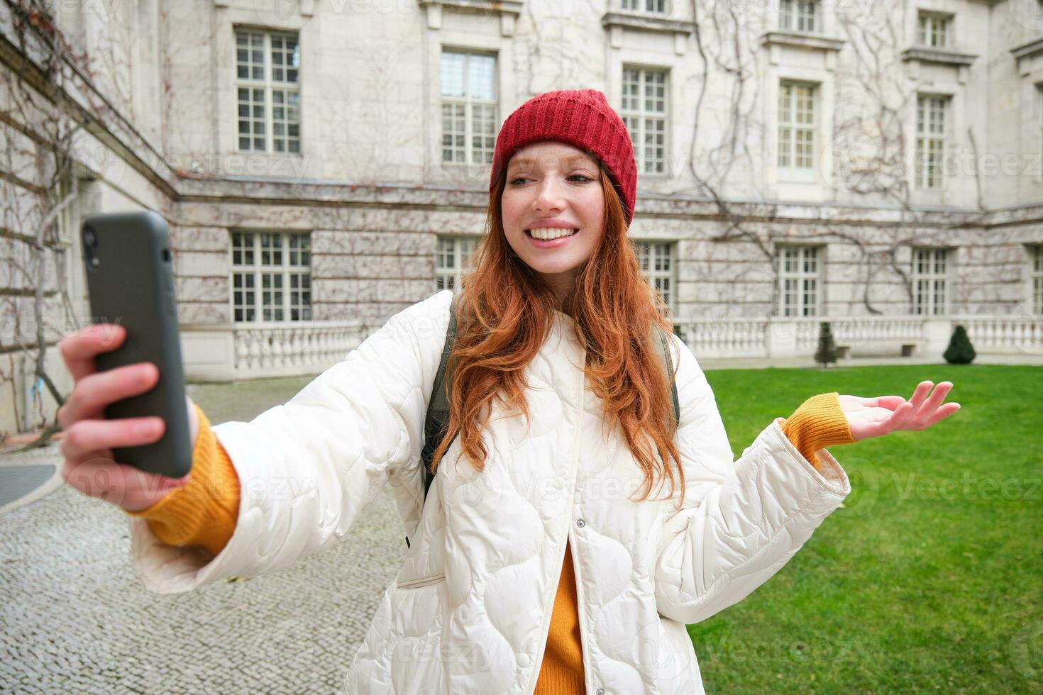 Portrait of happy tourist, girl with backpack, video chats, shows building, sightseeing attraction to friend, poses near landmark and smiles photo