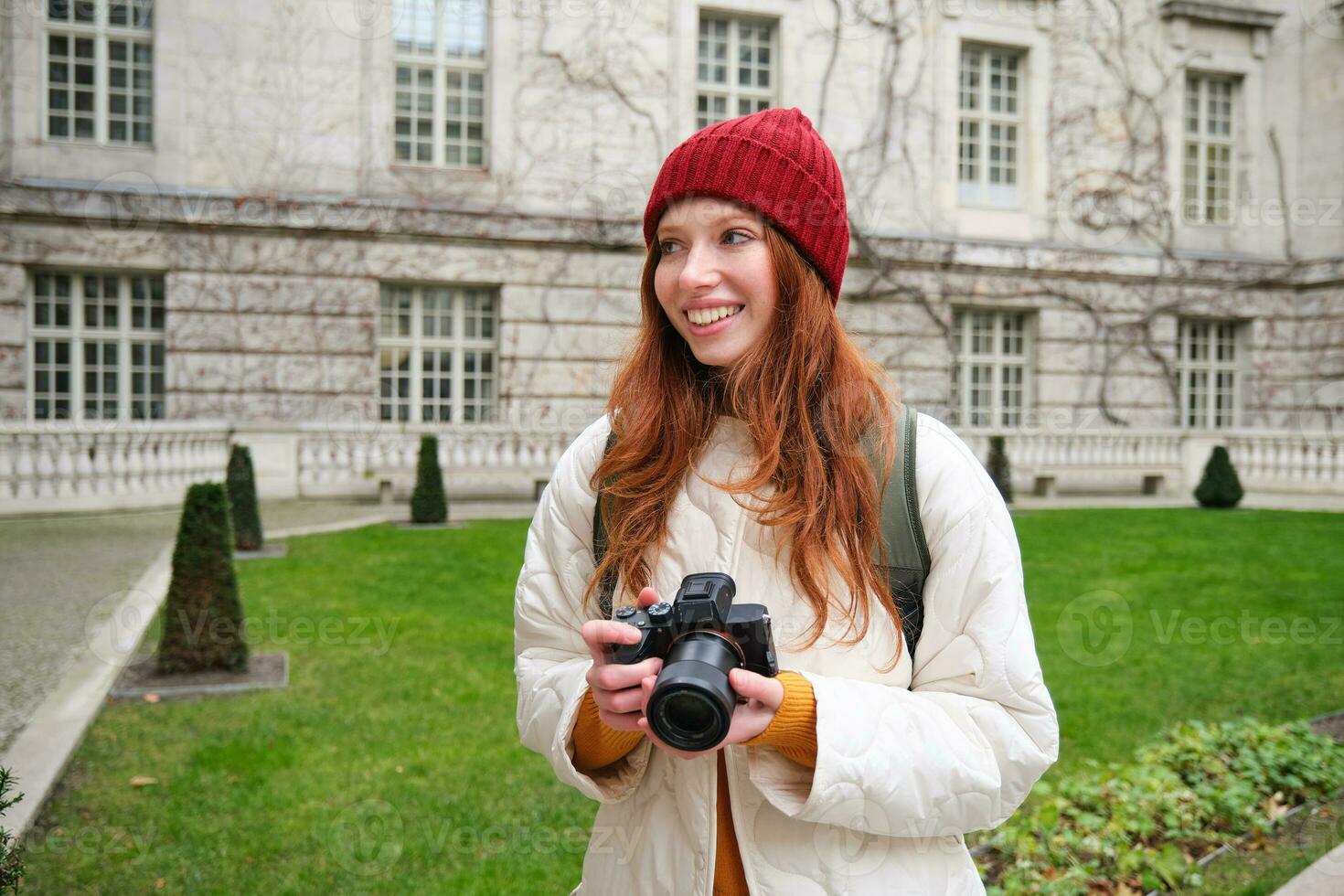 Redhead girl photographer takes photos on professional camera outdoors, captures streetstyle shots, looks excited while taking pictures