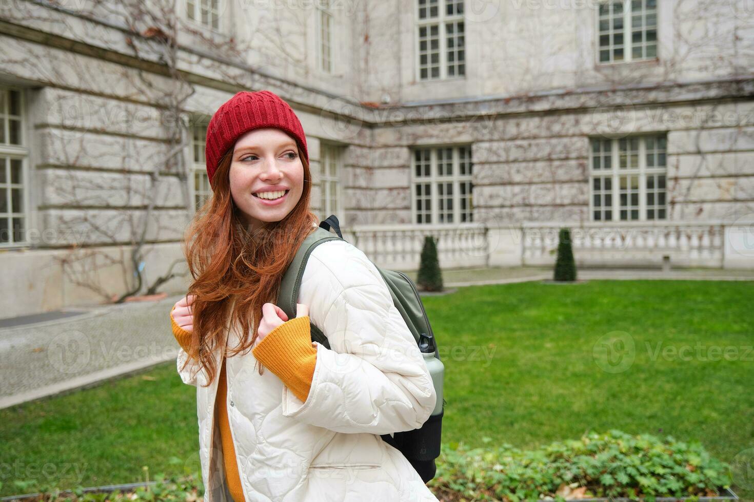 Beautiful redhead woman with backpack, tourist walking around city park and looks around, wears red hat and warm coat photo