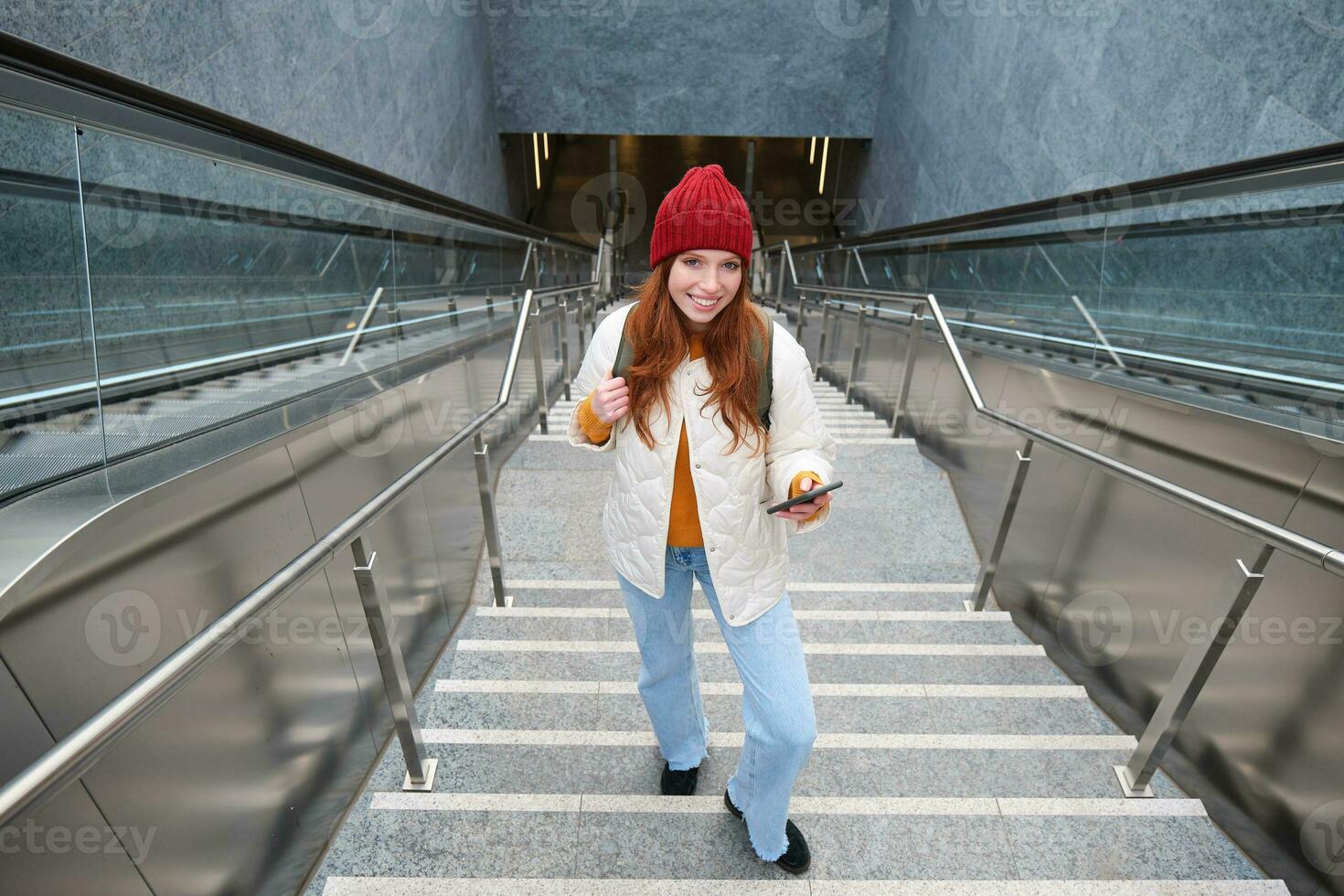 Outdoor shot of young woman plans route, follows map on smartphone app, goes up stairs with backpack and smiles photo