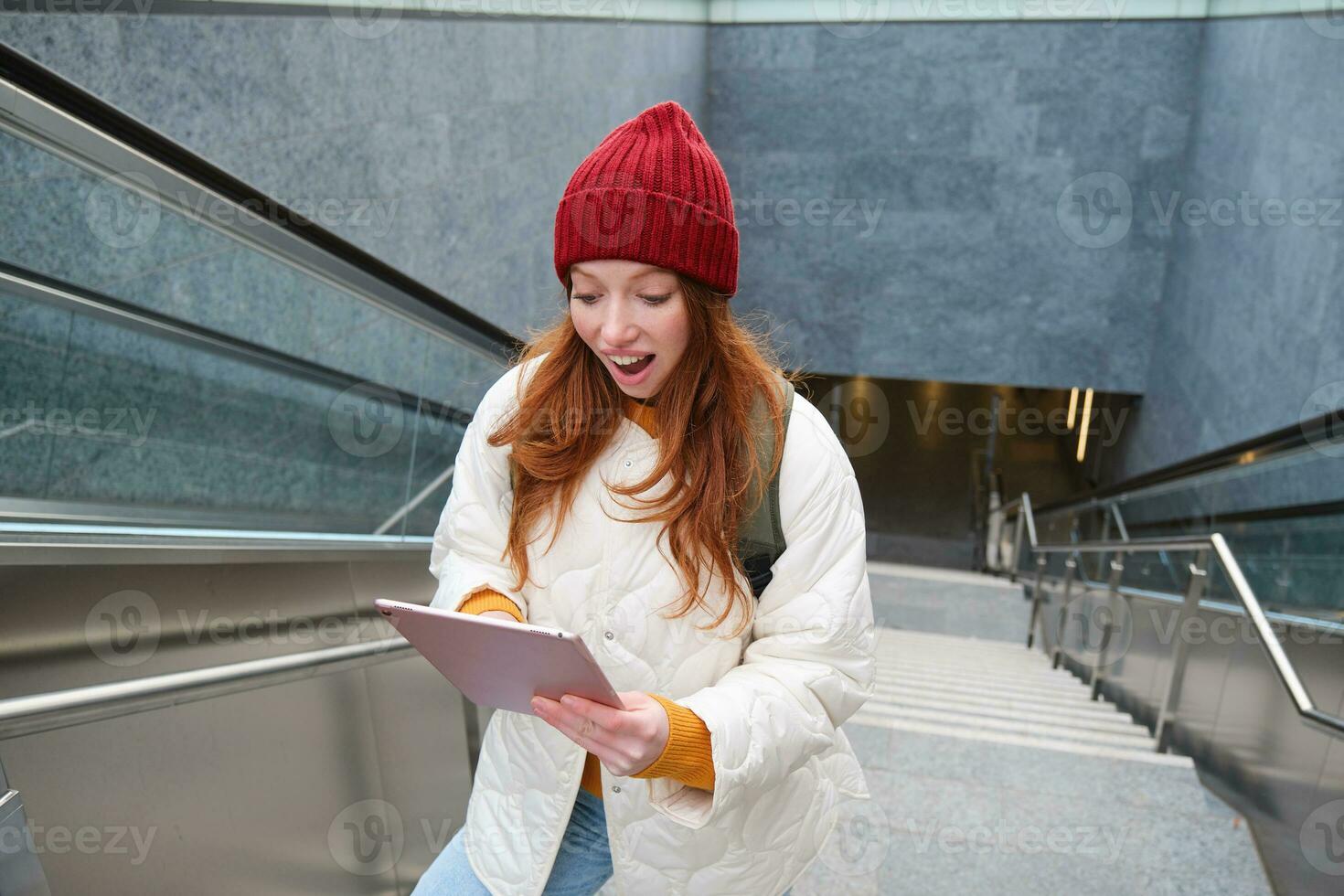 Portrait of girl student, walks up stairs from subway and looks at digital tablet with shocked, surprised face expression, reading amazing news photo