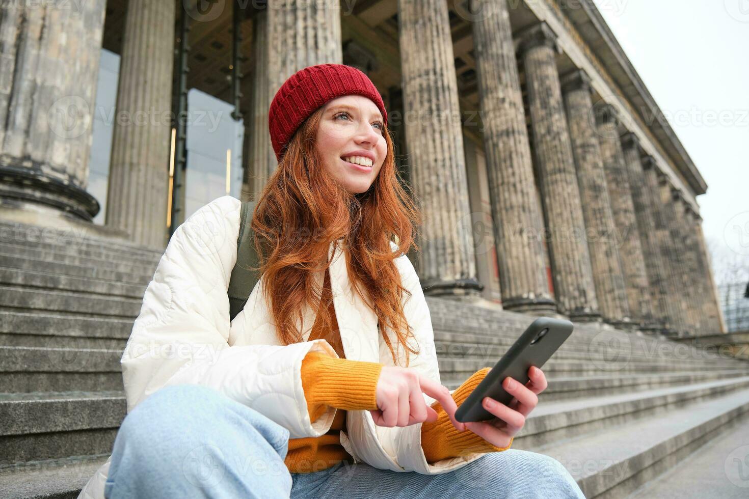 Portrait of young urban girl in red hat, sits on stairs near museum, holds mobile phone, connects to public wifi and surfs net, uses smartphone apps photo